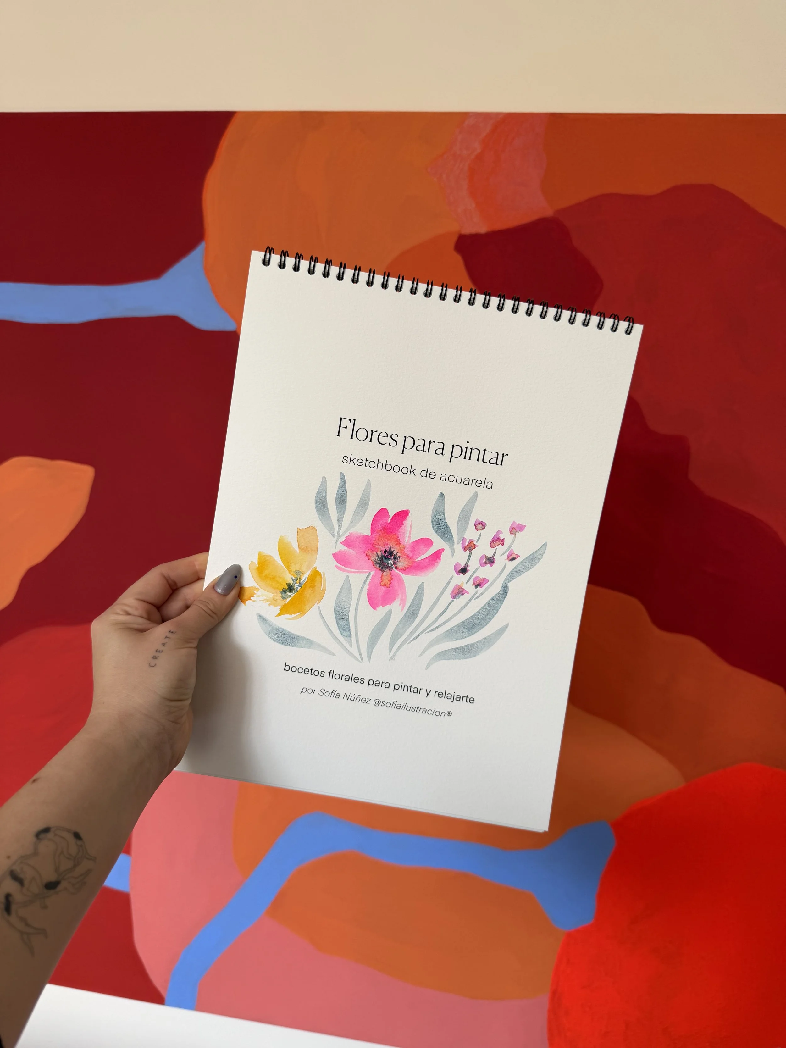 Libro titulado 'Flores para pintar, sketchbook de acuarela' con portada ilustrada de flores en acuarela, sostenido por una mano con tatuajes, frente a un fondo de pintura abstracta con colores cálidos y líneas azules.