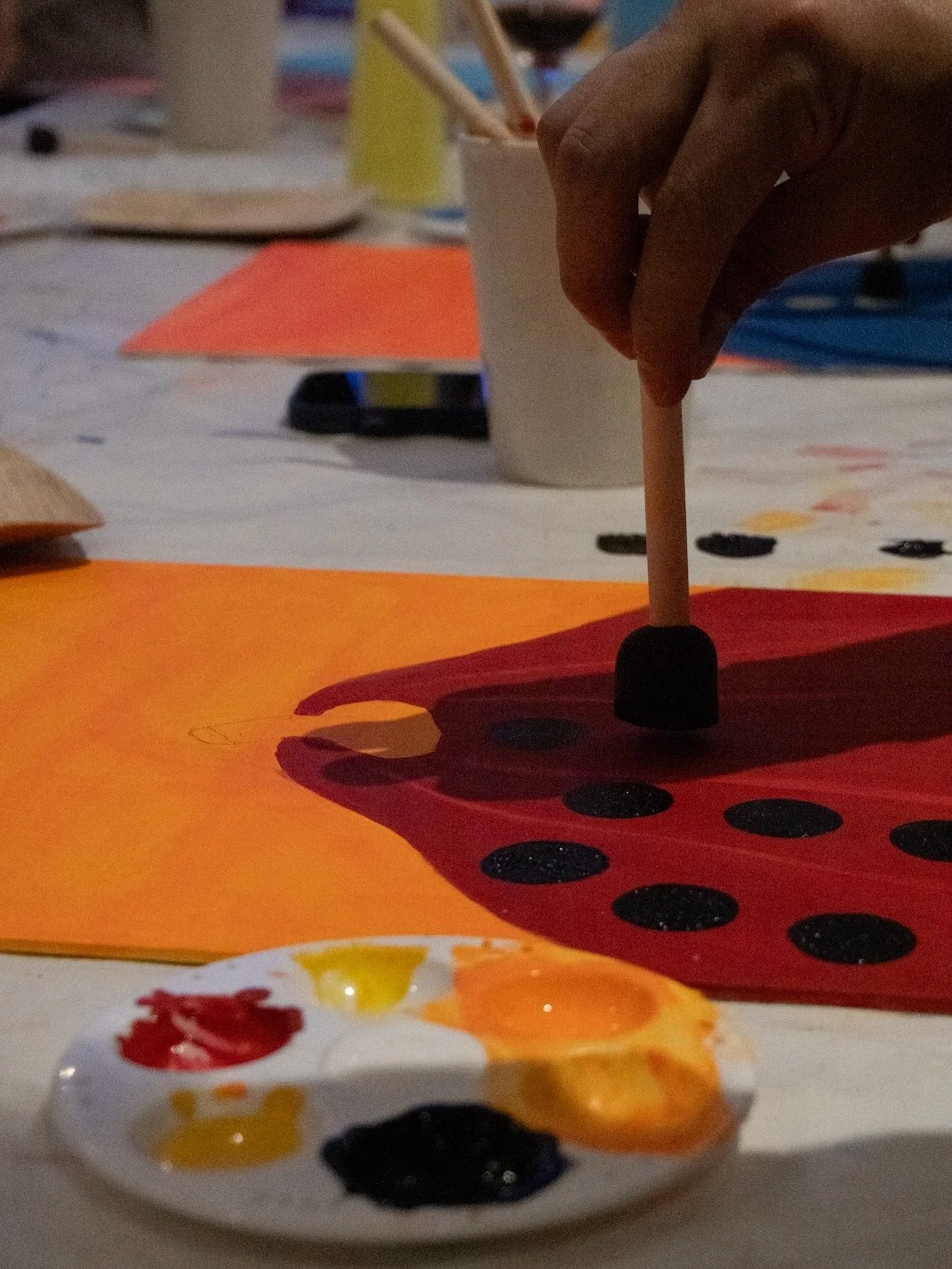 Mano colocando un palillo en una figura de papel pintada de rojo con puntos negros, sobre fondo naranja. Mesa con otros papeles y pintura.