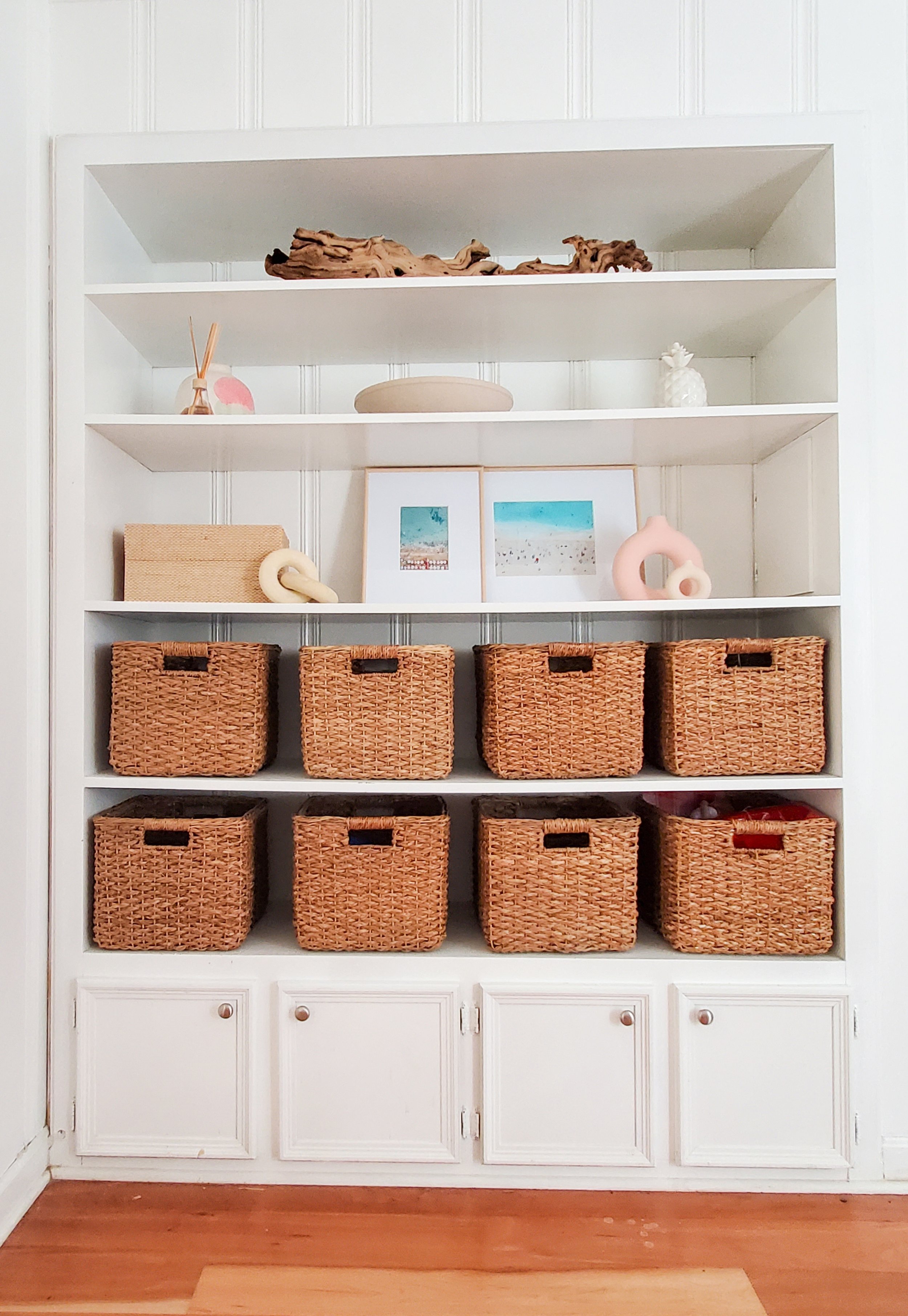 Playroom Decorated & Organized by Interior Designer Samantha Leonard. There is a large white built-in featuring shelves and small cabinets. There is coastal decor on the top three shelves with two rows of hyacinth baskets to contain toys