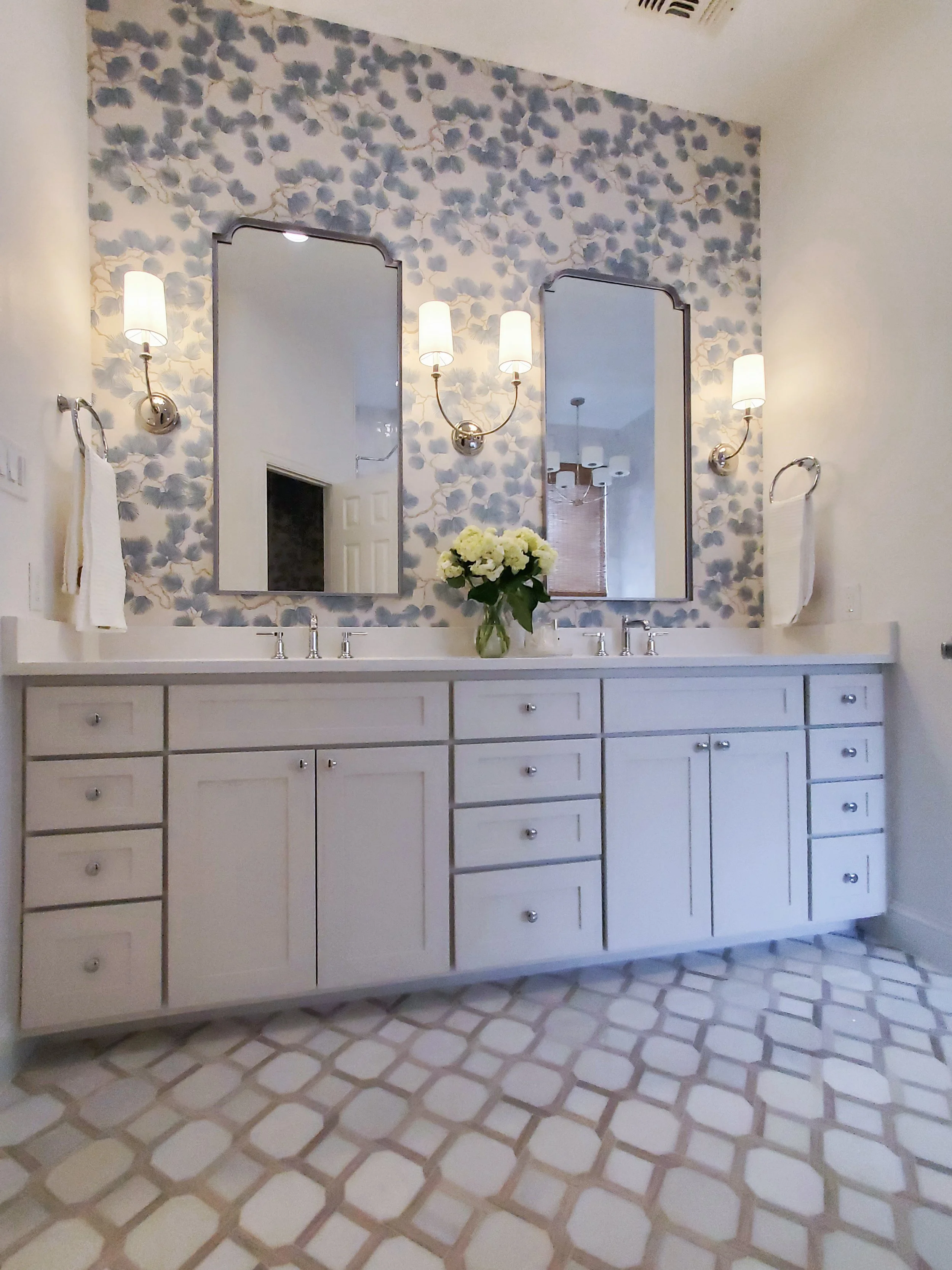 Double vanity with mirrors, sinks, and polished chrome wall sconces with blue floral wallpaper in the background in a Primary Bathroom in McKinney, Texas. Interior Design by Samantha Leonard Designs