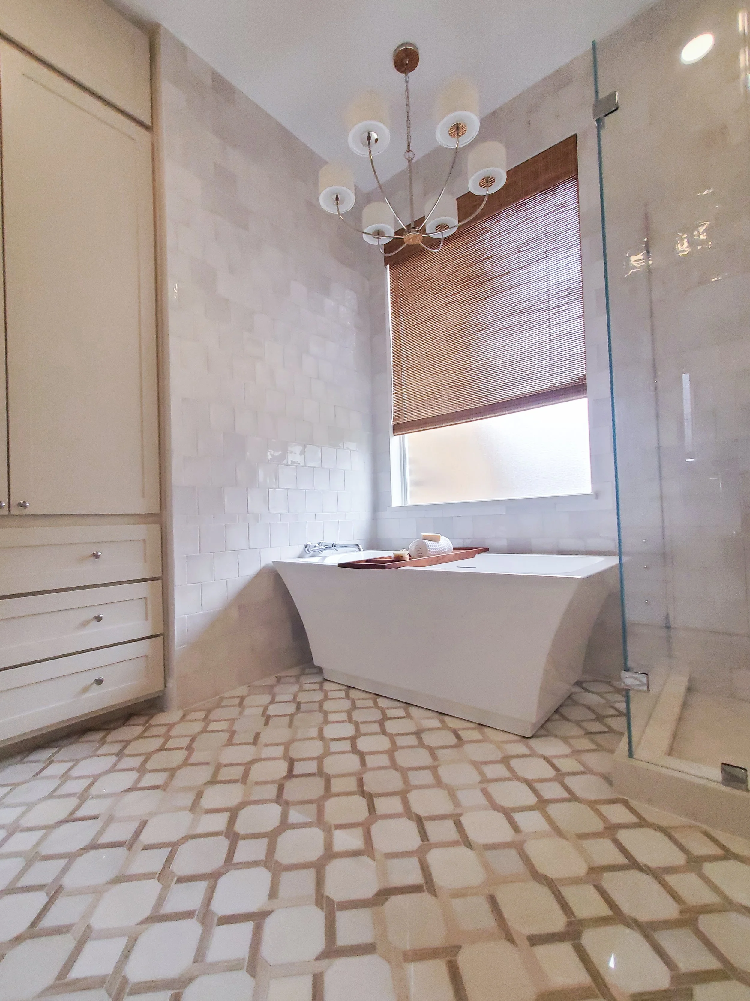 A white freestanding tub with a polished nickel chandelier above it on a patterned floor tile in a Primary Bathroom in McKinney, Texas. Interior Design by Samantha Leonard Designs