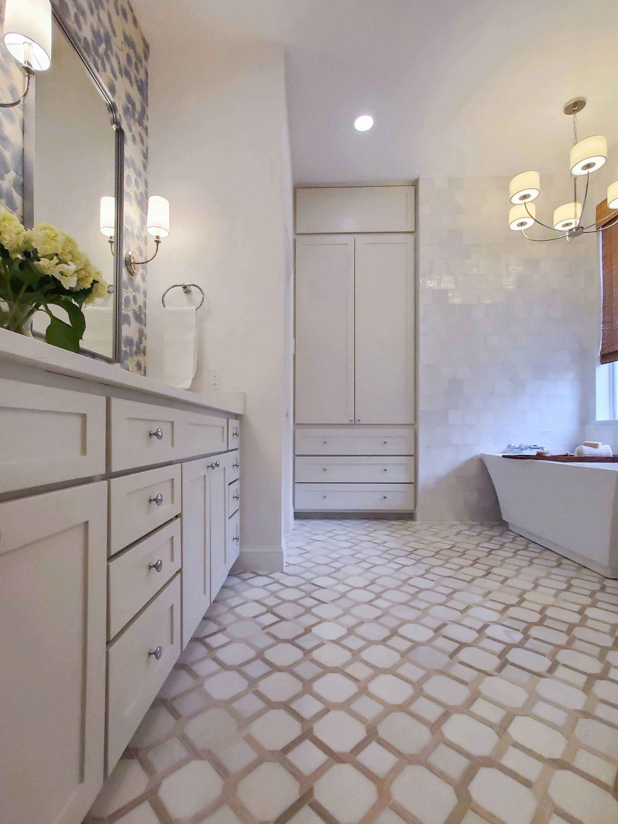 A white freestanding tub with a polished nickel chandelier above it on a patterned floor tile in a Primary Bathroom in with a cream colored double vanity on the side in McKinney, Texas. Interior Design by Samantha Leonard Designs