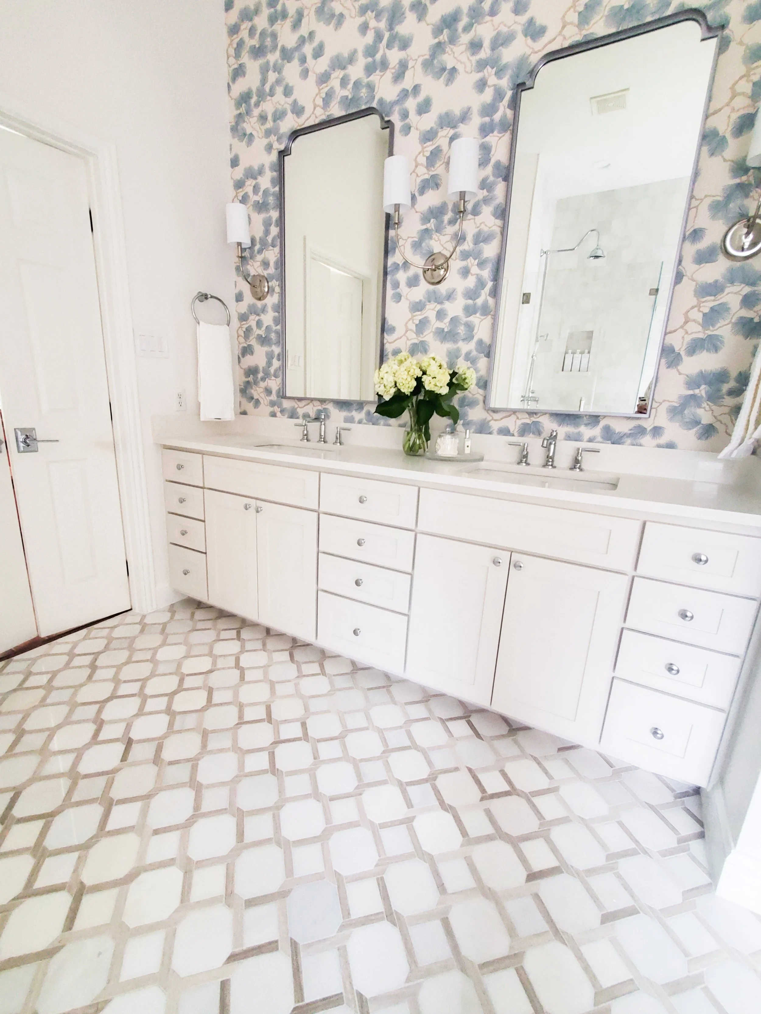Double vanity with mirrors, sinks, and polished chrome wall sconces with blue floral wallpaper in the background in a Primary Bathroom in McKinney, Texas. Interior Design by Samantha Leonard Designs