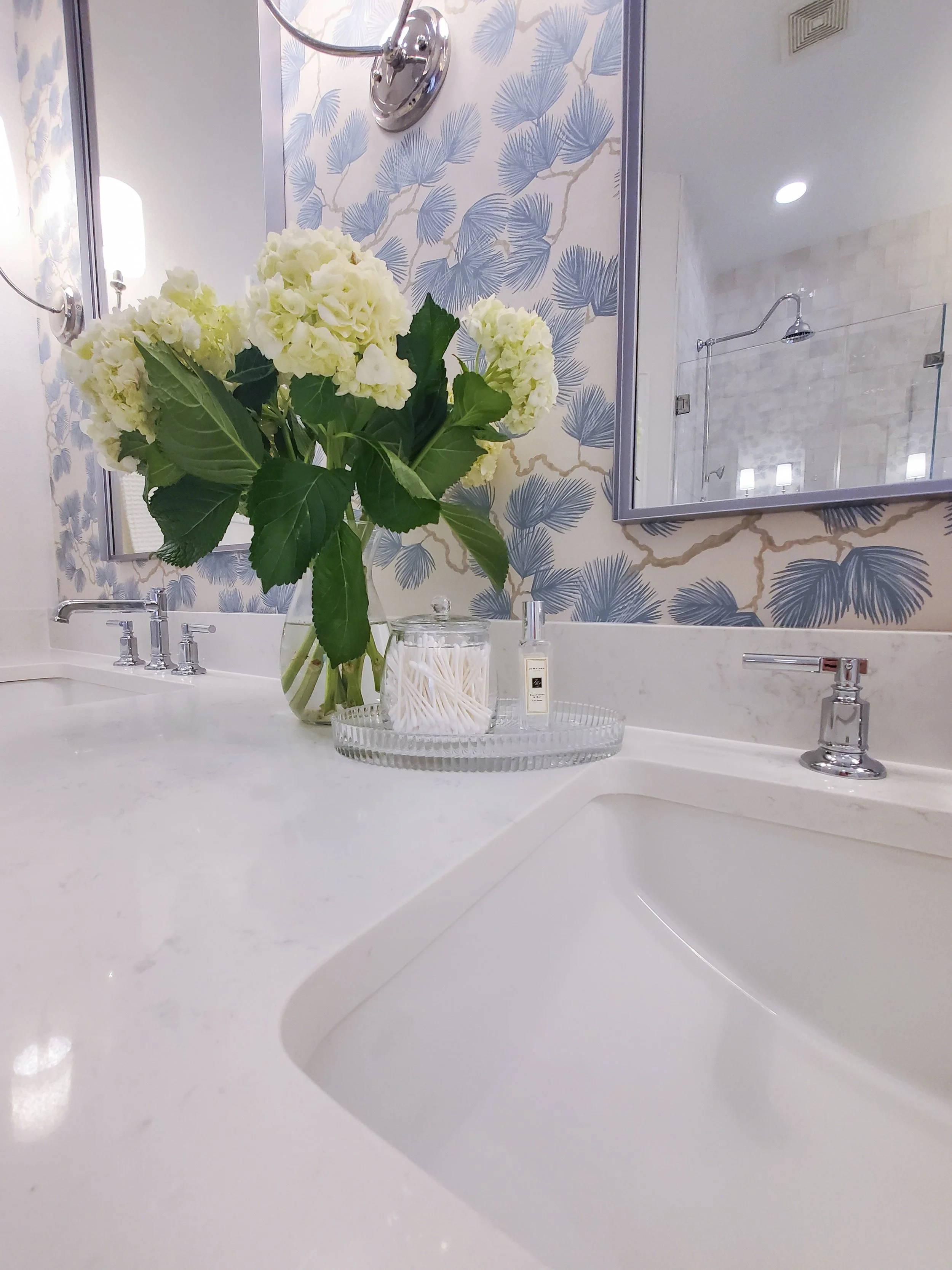 A close up of a quartz countertop with sink and polished chrome faucets and Blue Wispy Flower Wallpaper in a Primary Bathroom in McKinney, Texas. Interior Design by Samantha Leonard Designs
