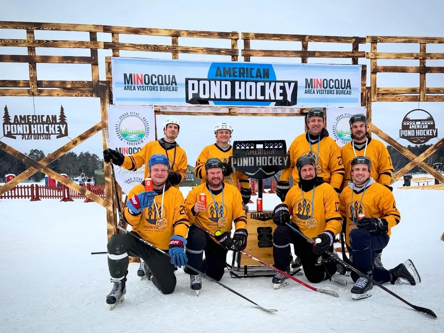 Rocky Reef Brewing Company pond hockey team celebrating their Open A championship at American Pond Hockey 2026 in front of the APH arch and championship torch on Lake Minocqua in Wisconsin