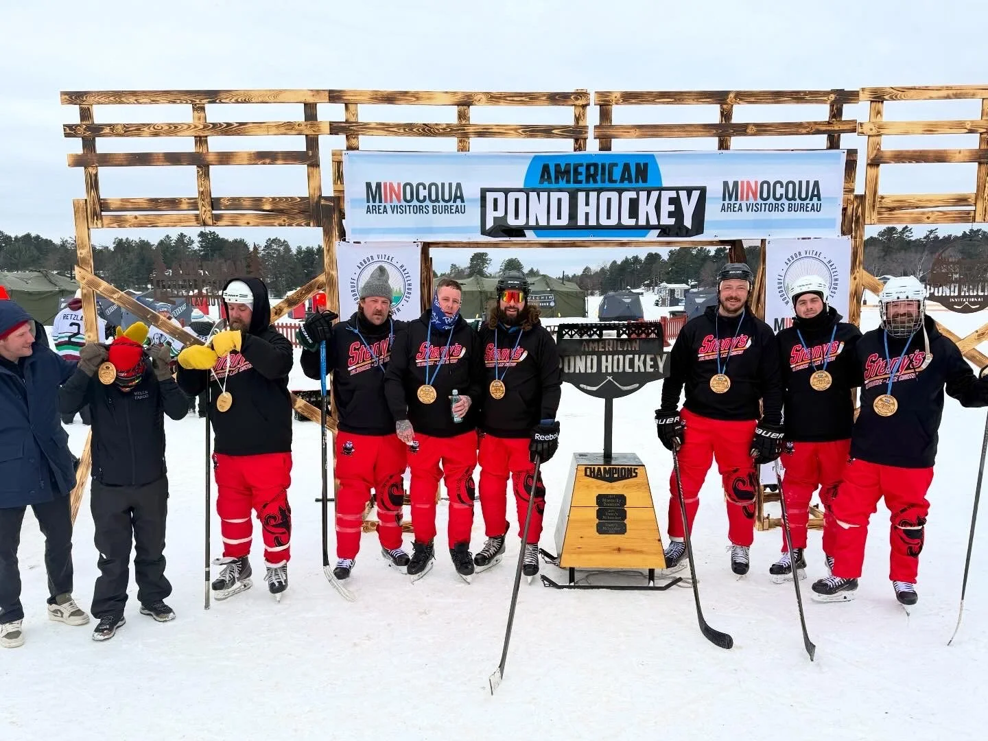 Steny's Milwaukee celebrating their 2026 American Pond Hockey championship in teh Open B Division on Lake Minocqua, with a backdrop of the APH bridge entryway and the Championship torch. Each player is wearing their wooden first place medals