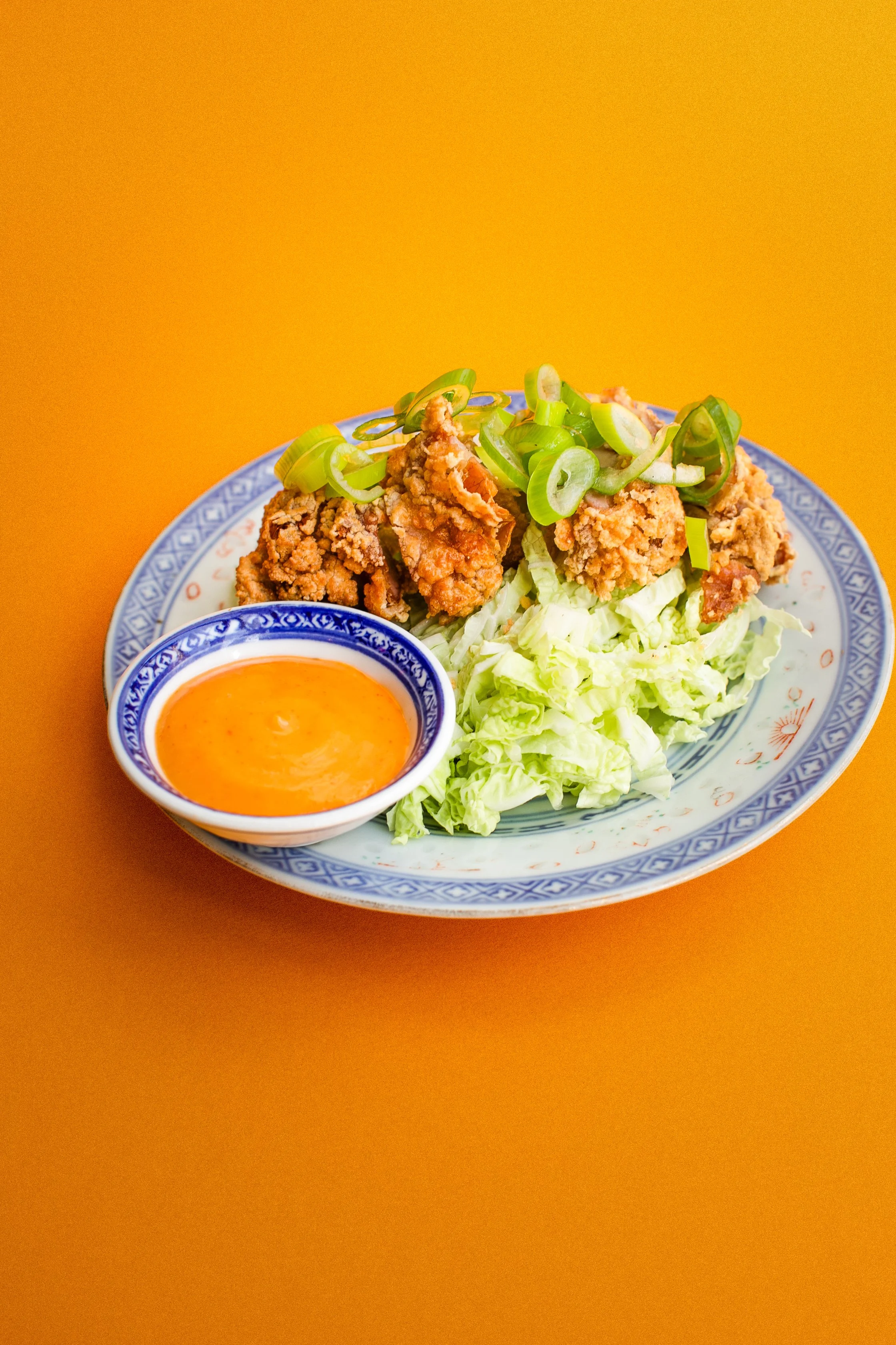 Assiette de poulet frit avec sauce à côté, garnie de ciboule et de laitue, sur une table orange.
