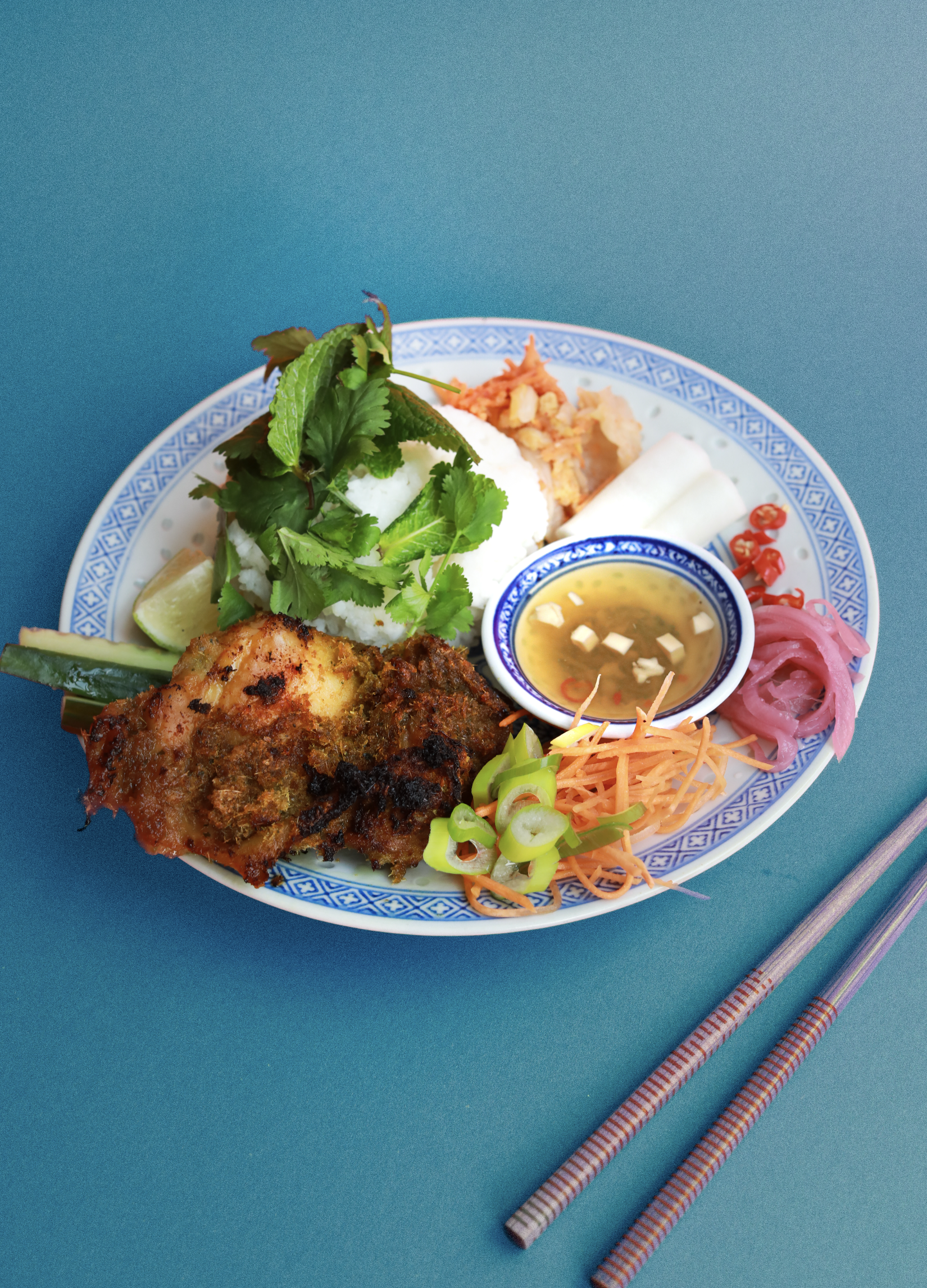 Assiette de cuisine vietnamienne avec du riz, du poulet mariné, des herbes fraîches, des légumes, une sauce, et des garnitures colorées, accompagnée de baguettes.