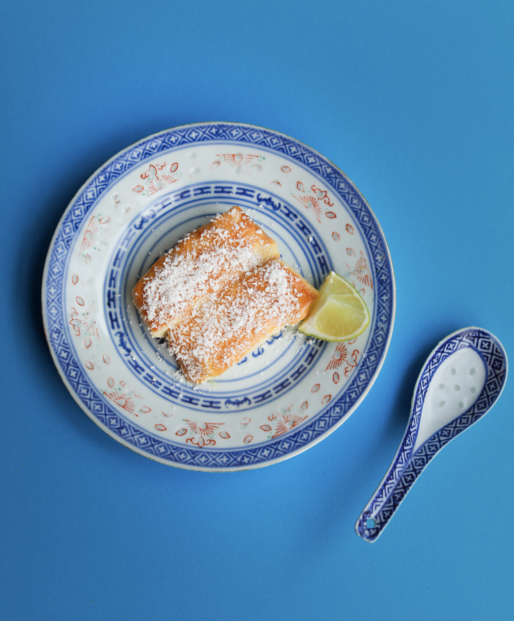Un dessert à la mangue, avec une tranche de citron vert, présenté dans une assiette en porcelaine décorée de motifs bleus et rouges. Deux cuillères en porcelaine assorties reposent à côté, le tout sur un fond bleu uni.