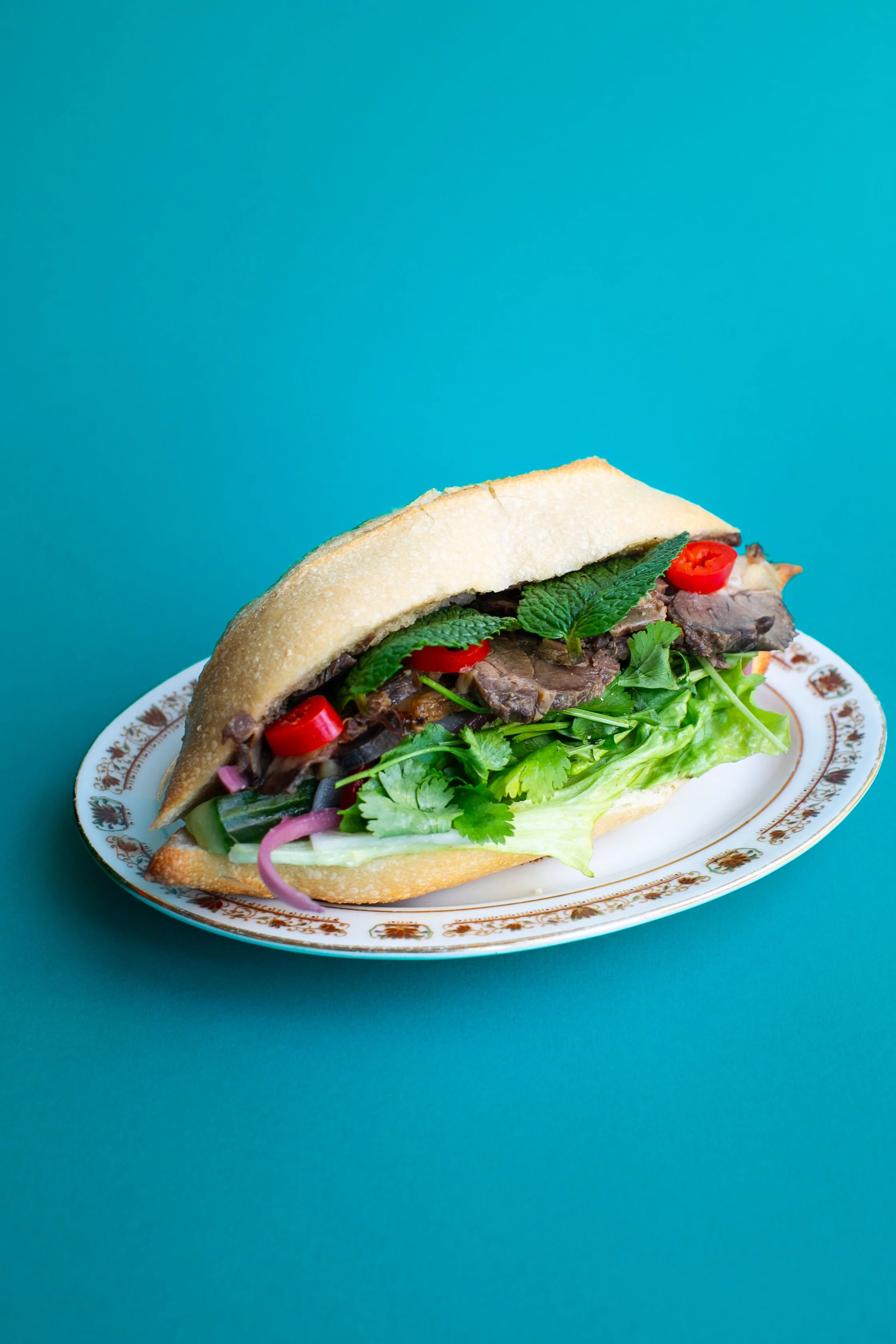Sandwich vietnamien avec légumes frais et viande sur une assiette décorative, posé sur un fond bleu turquoise.