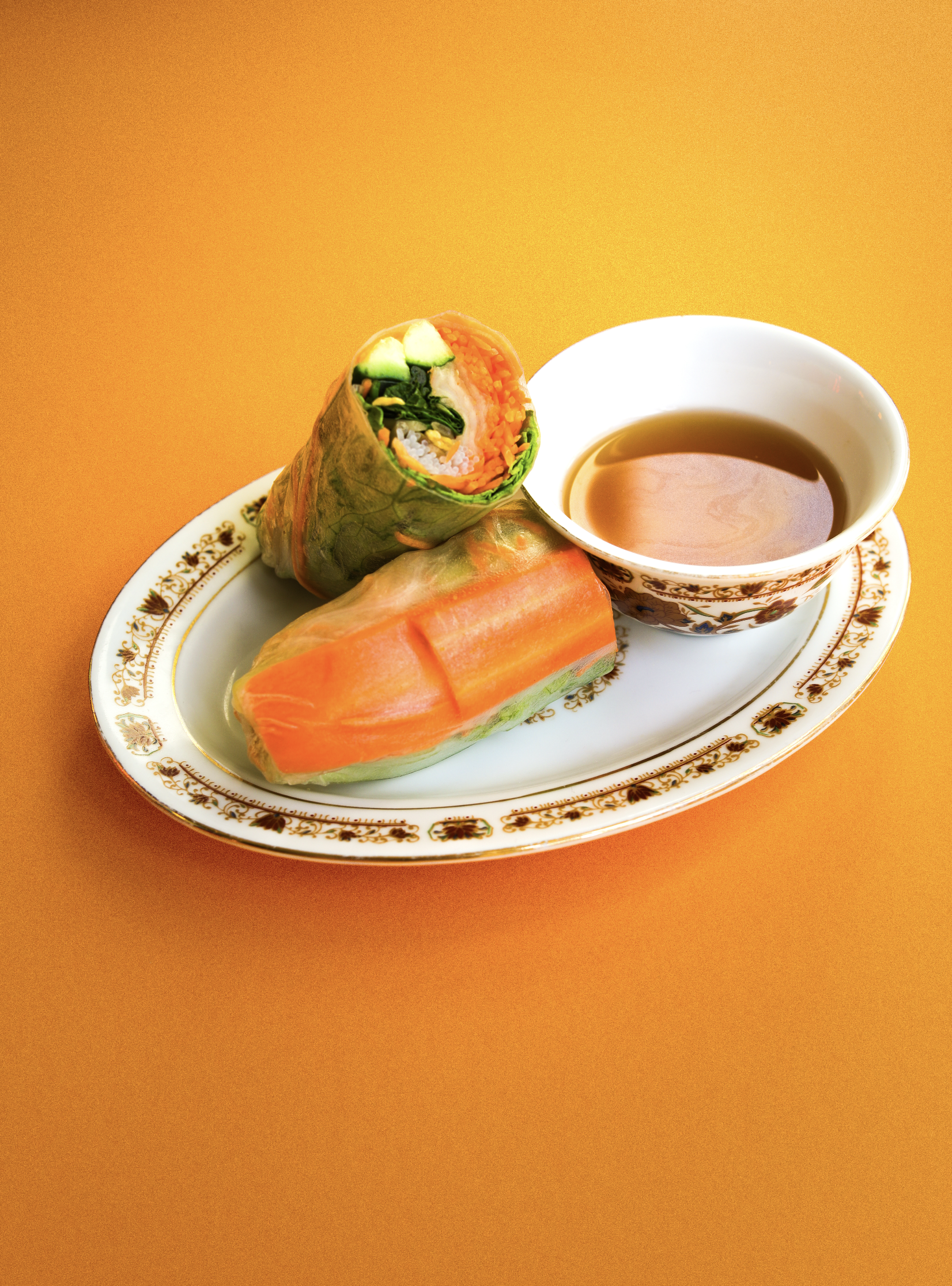 Rouleau de printemps avec légumes, morceau de saumon fumé, sauce dans un bol avec décor floral, sur une assiette ornée, fond orange.