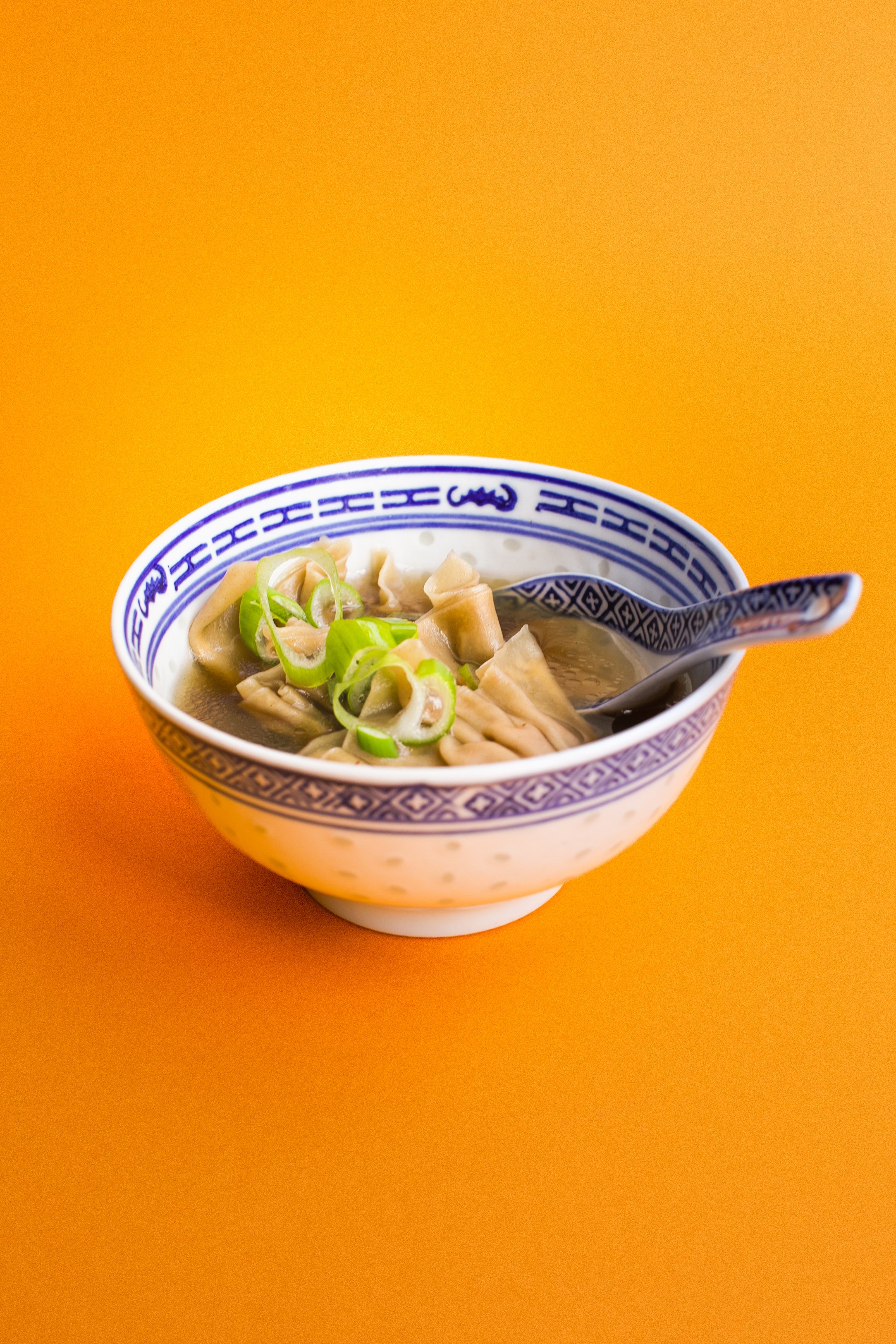 Bol de soupe avec des morceaux de poulet et des rondelles de céleri, accompagné d'une cuillère en céramique à motifs chinois, placé sur une surface orange.