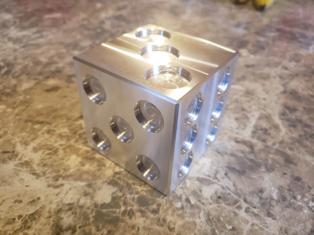 Metallic cube with circular indentations on each side, resembling a dice, placed on a marble surface.