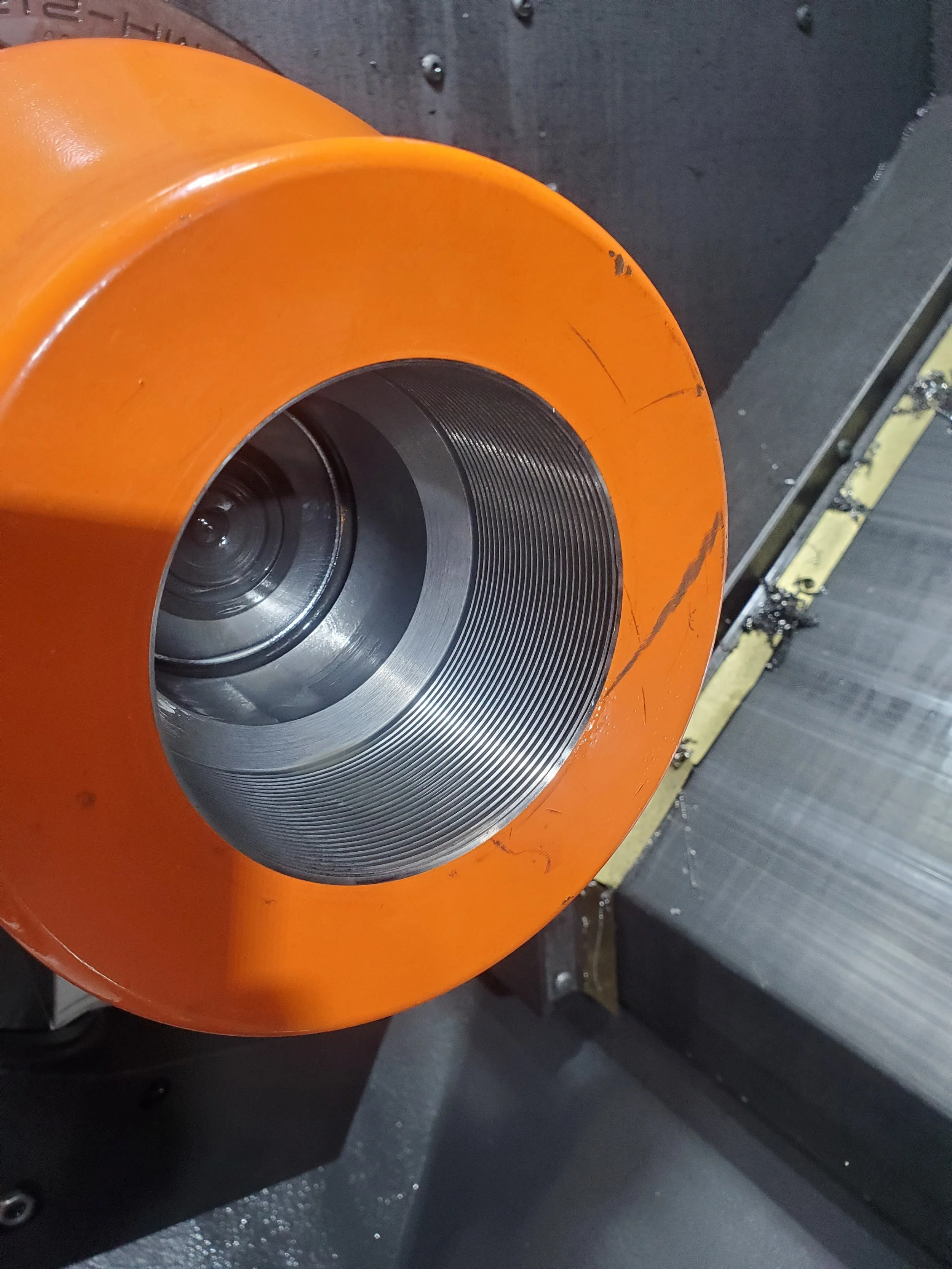 Close-up of an orange metal cylinder with an internal threaded surface, possibly part of industrial machinery, against a grey metallic background.