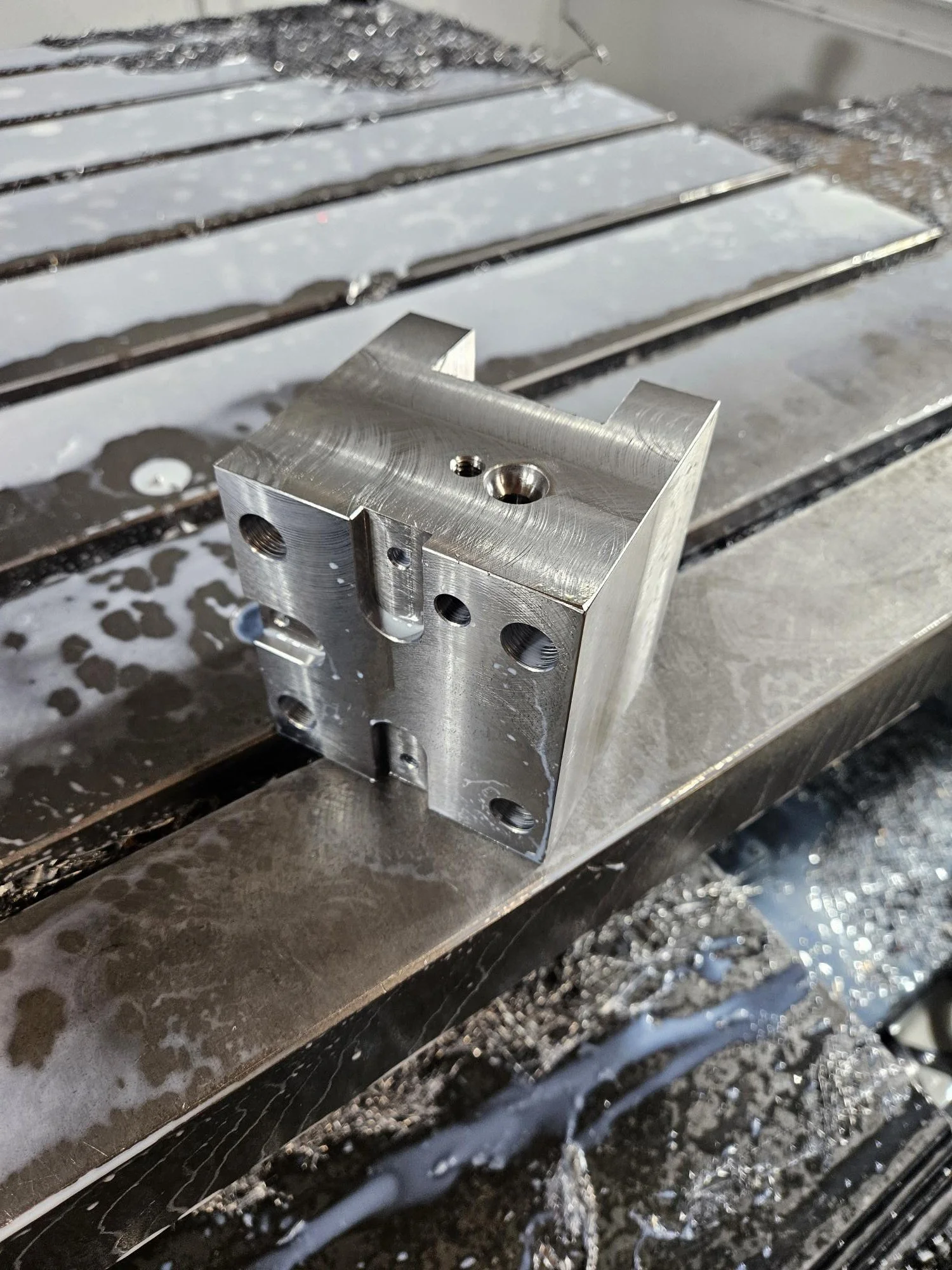 A metal block with multiple holes placed on a wet, metallic surface, possibly a machining table.