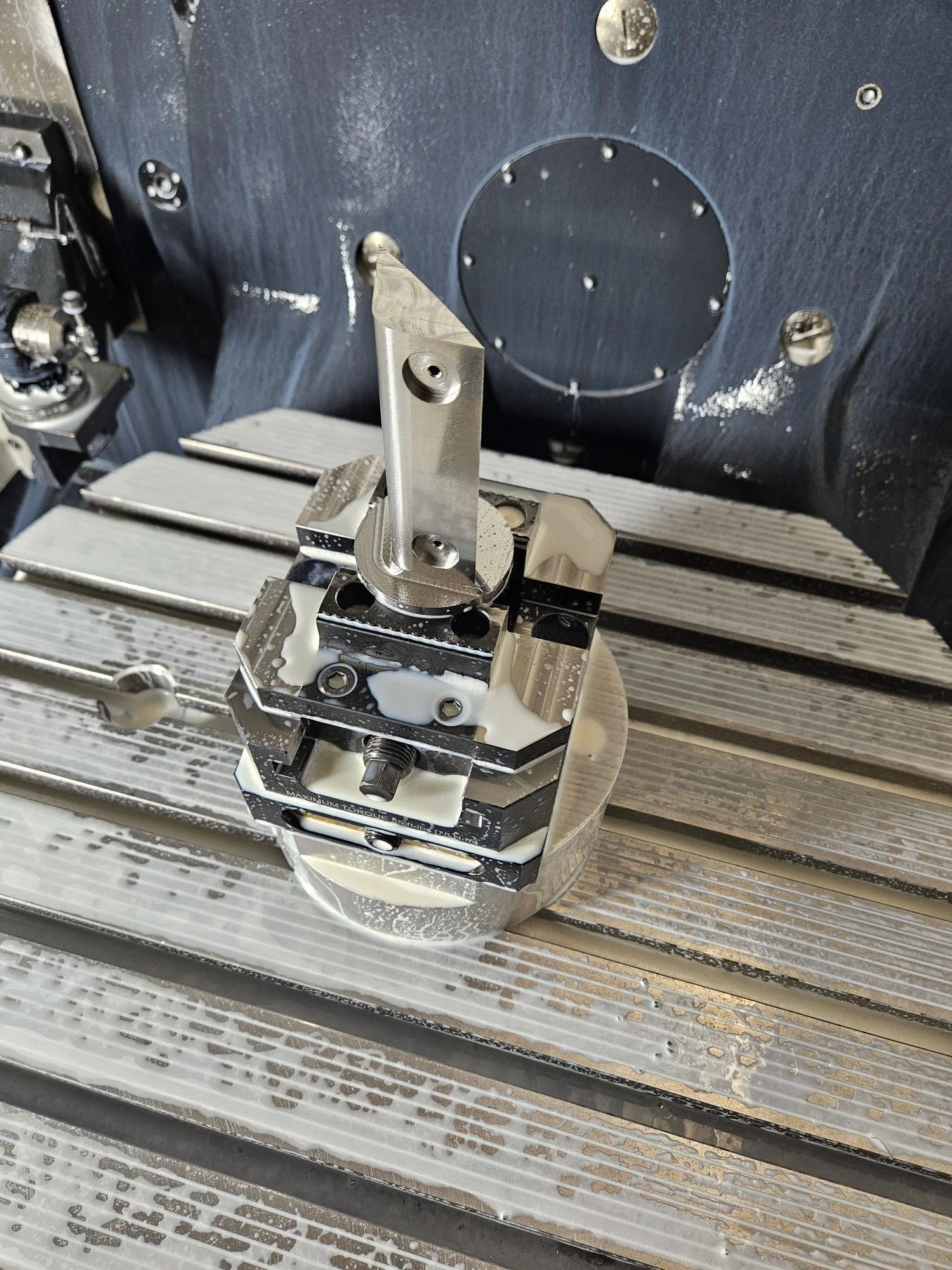 Metal part being machined, secured on a CNC milling machine with clamps, surrounded by metal swarf and coolant.