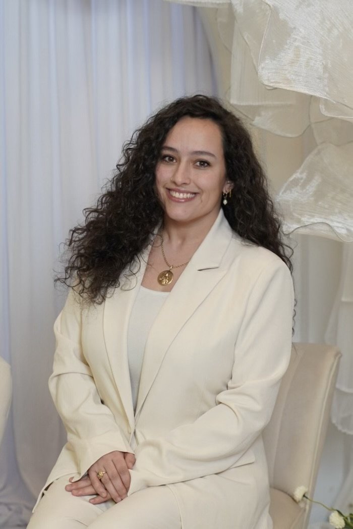 A woman with long, curly brown hair, wearing a white suit, sitting on a light-colored chair, smiling, with a light-colored background and sheer curtains.