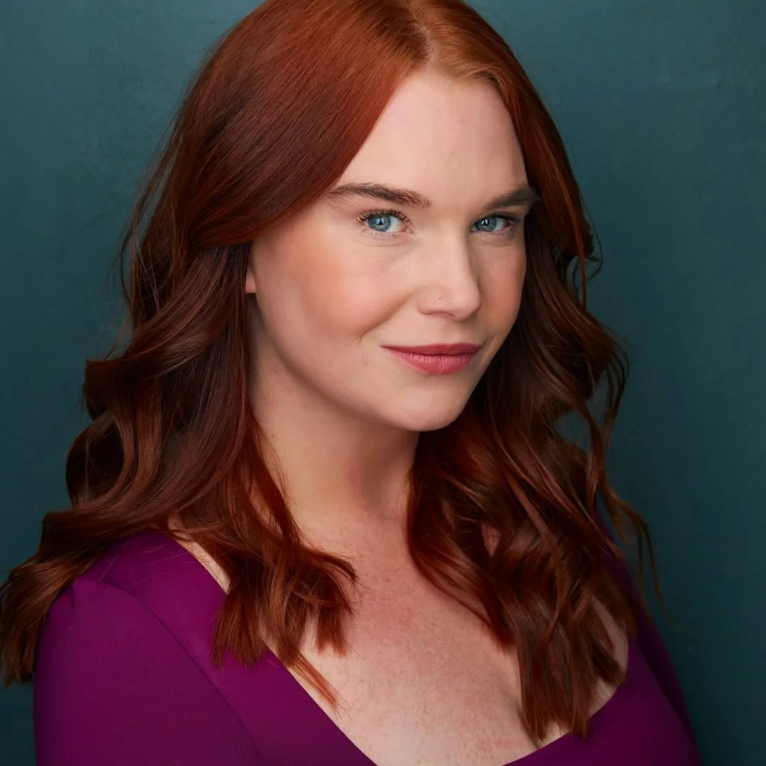 Close-up portrait of a woman with red hair and blue eyes, wearing a purple top, against a dark teal background.