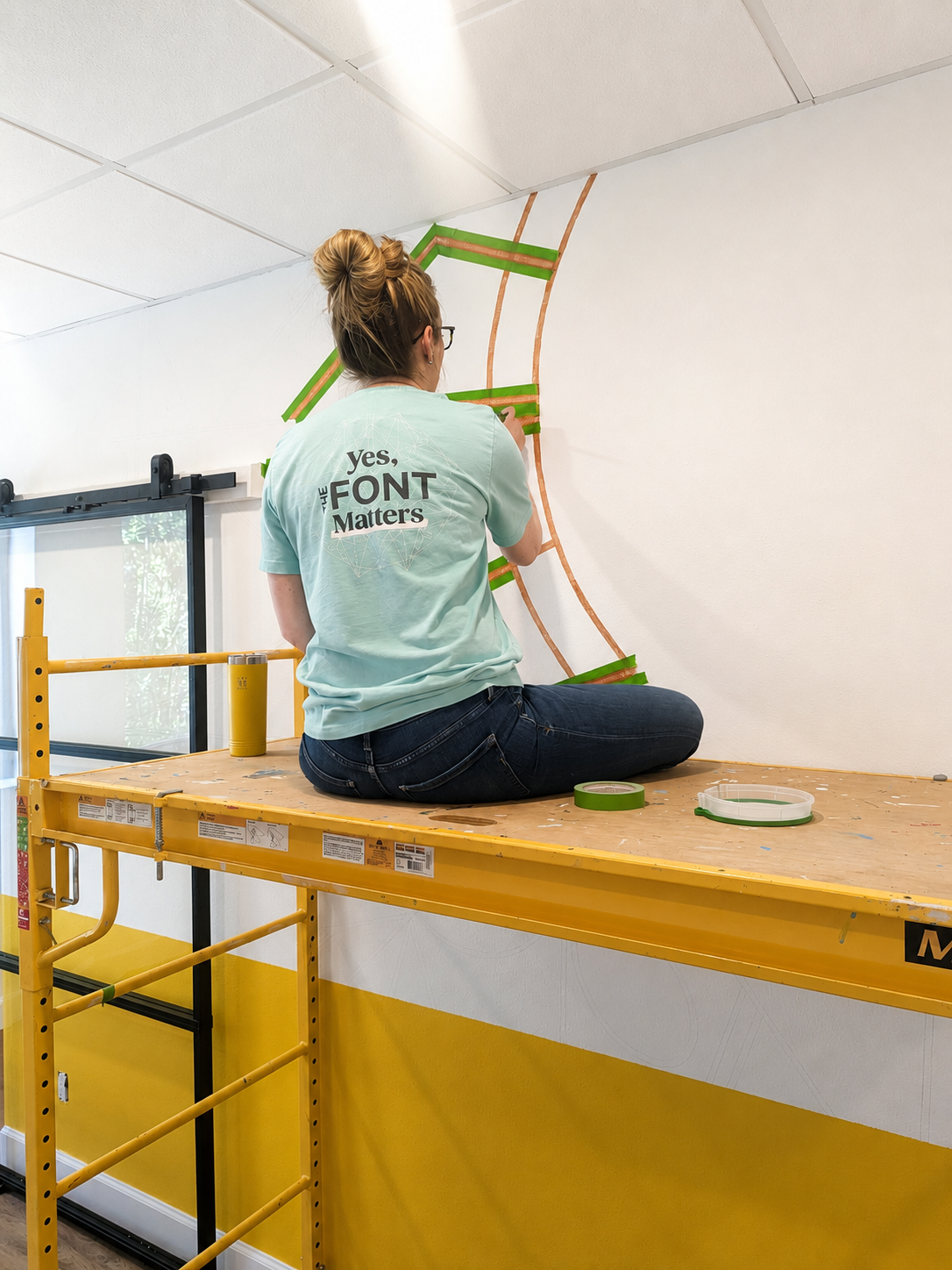 Heather sitting on top of scaffolding painting a mural in the Manifestive Design office