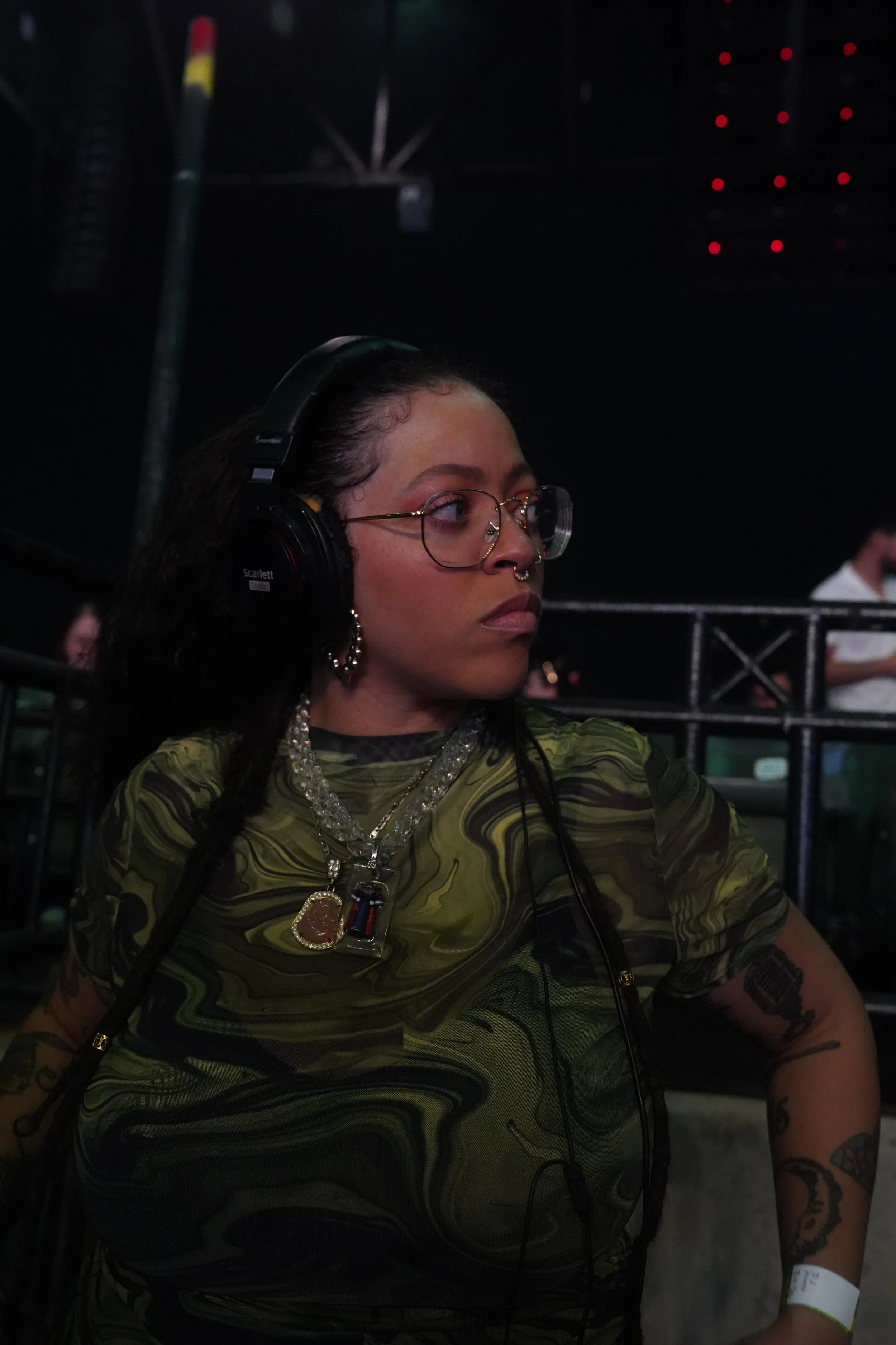 A woman with glasses, tattoos, and jewelry wearing a green and black patterned shirt and large headphones at a dark event.