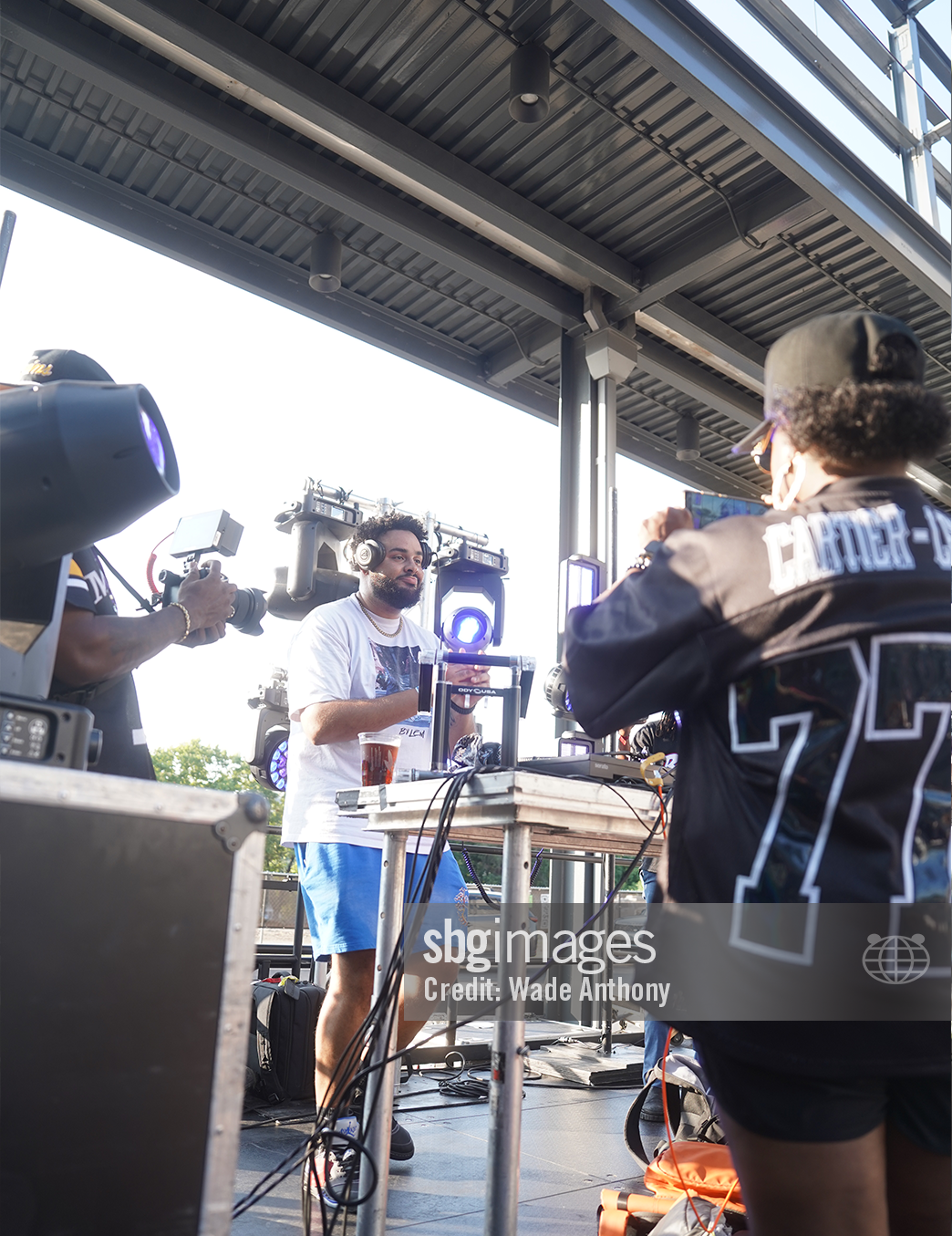 A DJ wearing headphones and a white t-shirt performs outdoors on a stage with lighting equipment, while a person in a black jersey with the number 77 records him with a smartphone. A cameraman films the scene. There are cables and equipment on the table in front of the DJ.