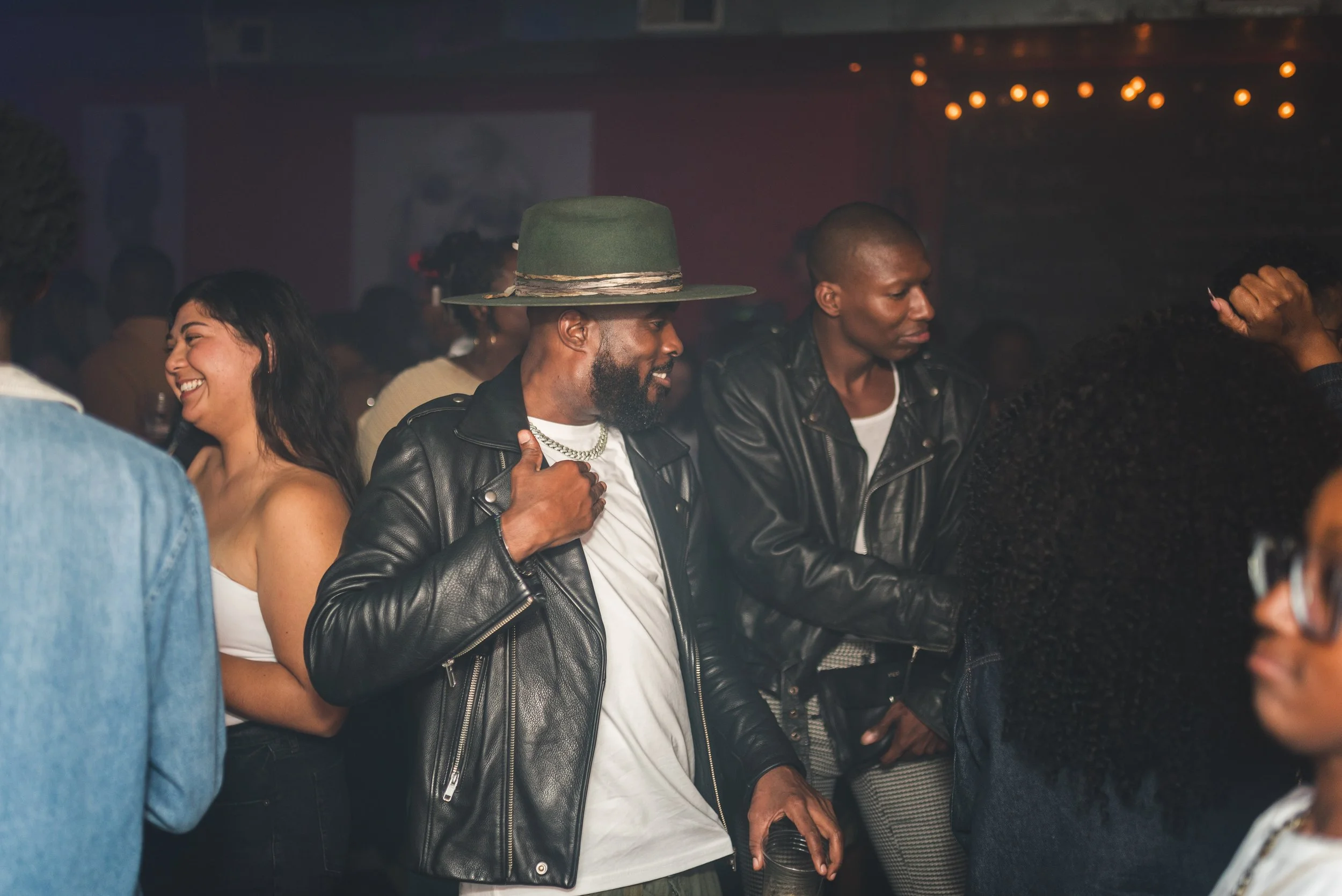 People socializing at a party or nightclub, with some dancing and smiling, in a dimly lit environment decorated with string lights.