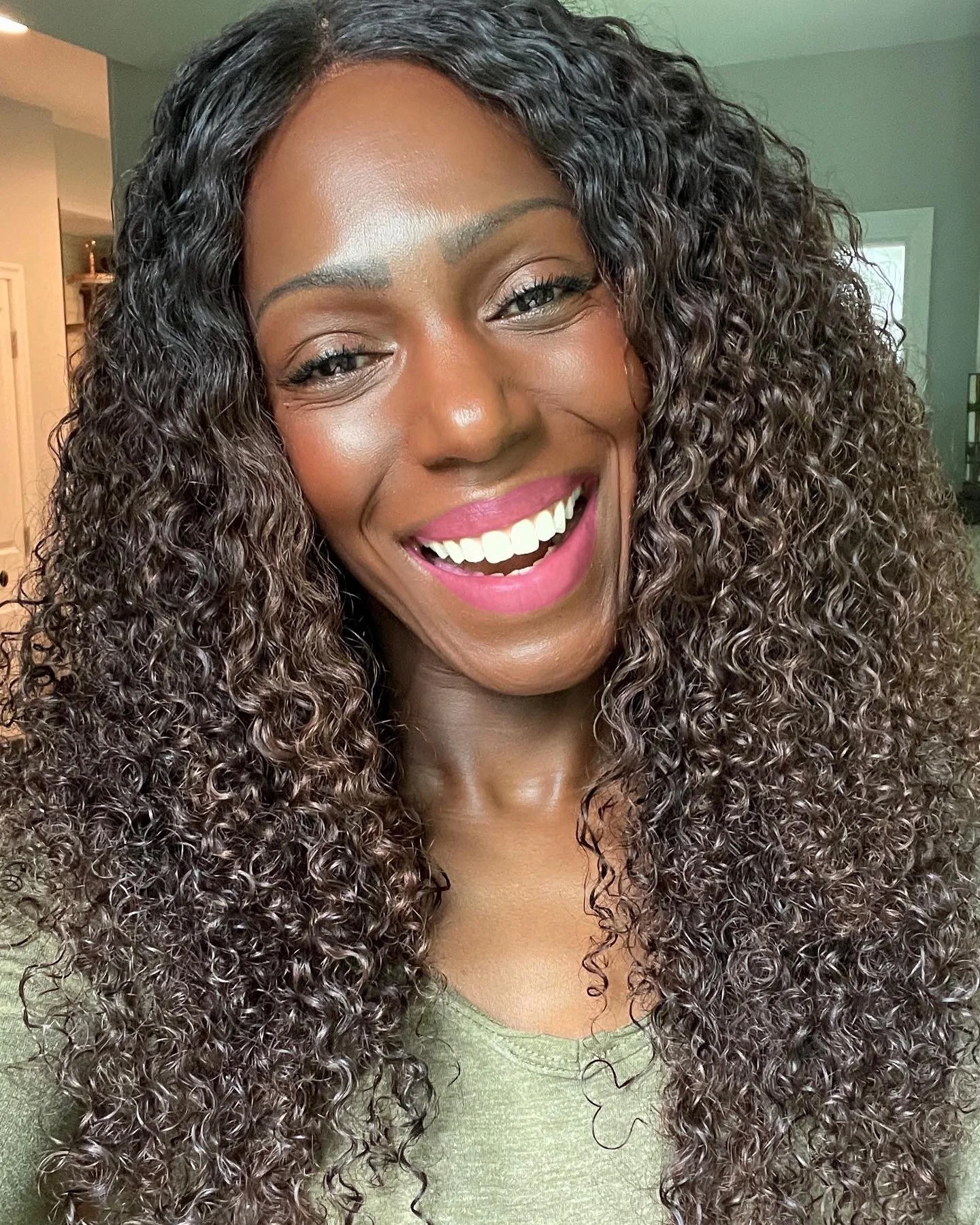 Smiling woman with long, curly black hair, wearing a black cut-out dress, standing in front of a light-colored textured background.