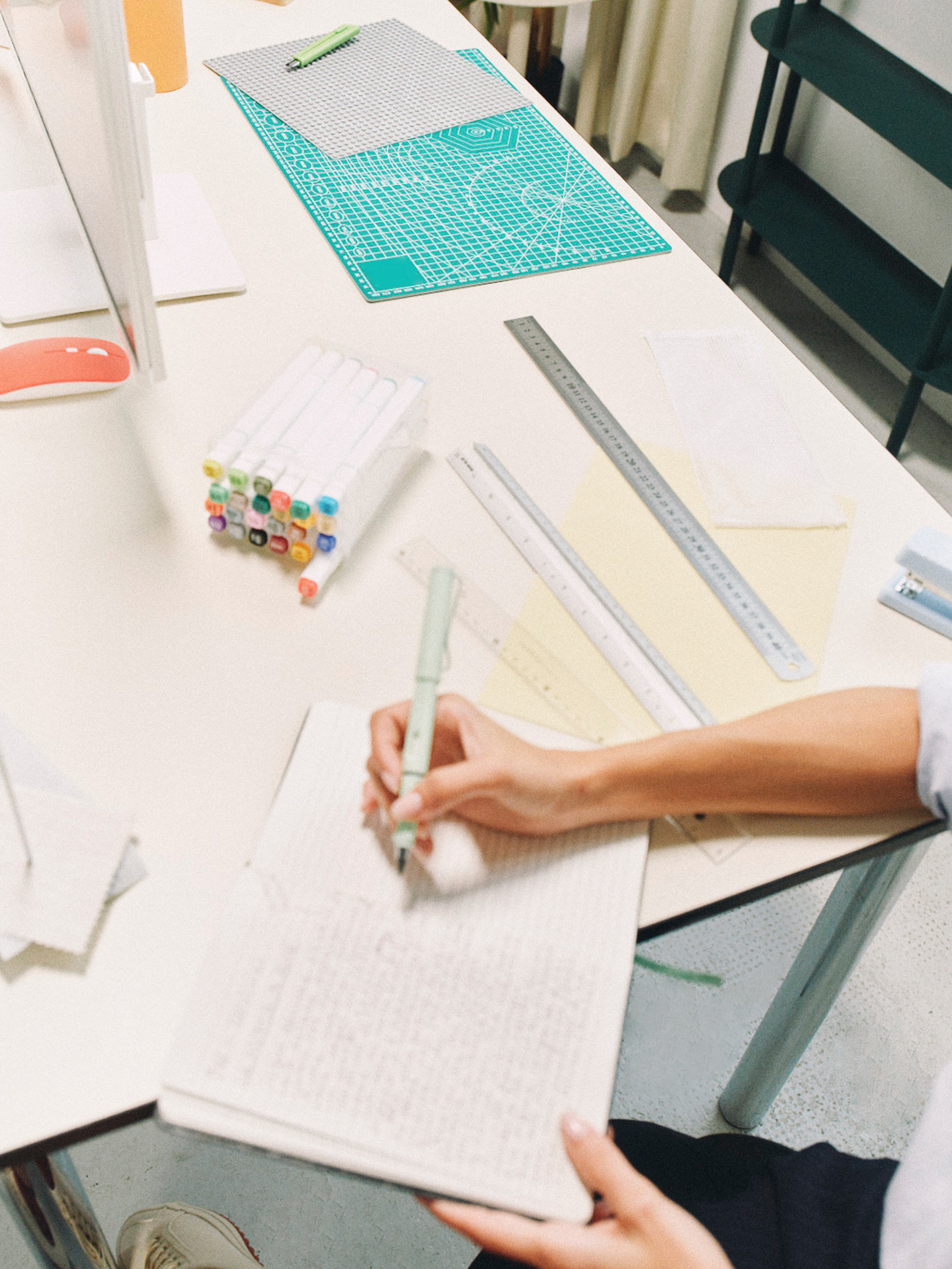 A person writing in a notebook at a table with drafting tools, colorful markers, a ruler, and various cutting mats.