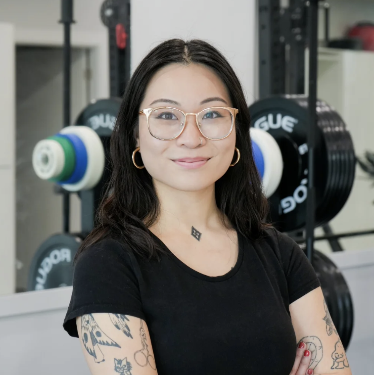 A woman with glasses and tattoos standing in a gym with weight plates behind her.