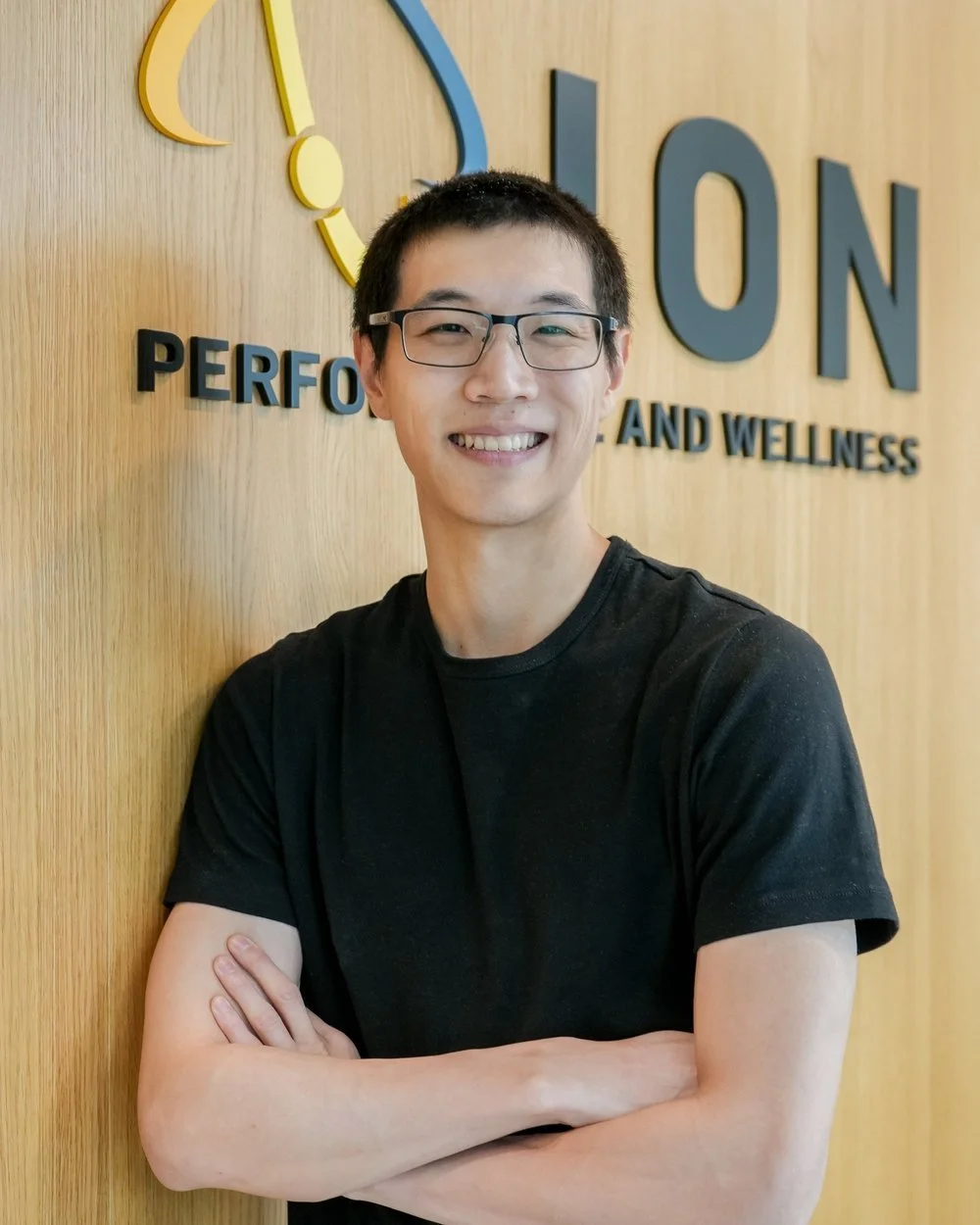 A young man with glasses smiling and crossing his arms, standing in front of a wooden wall with a logo that reads "ION PERFORMANCE AND WELLNESS."