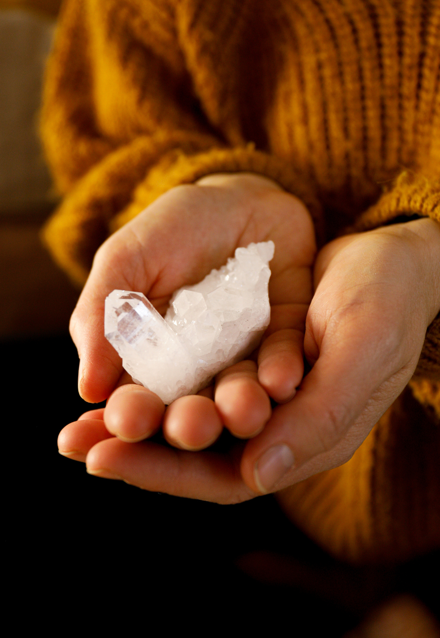 Person holding a rough, translucent white crystal in their hand, wearing a rust-colored knitted sweater.