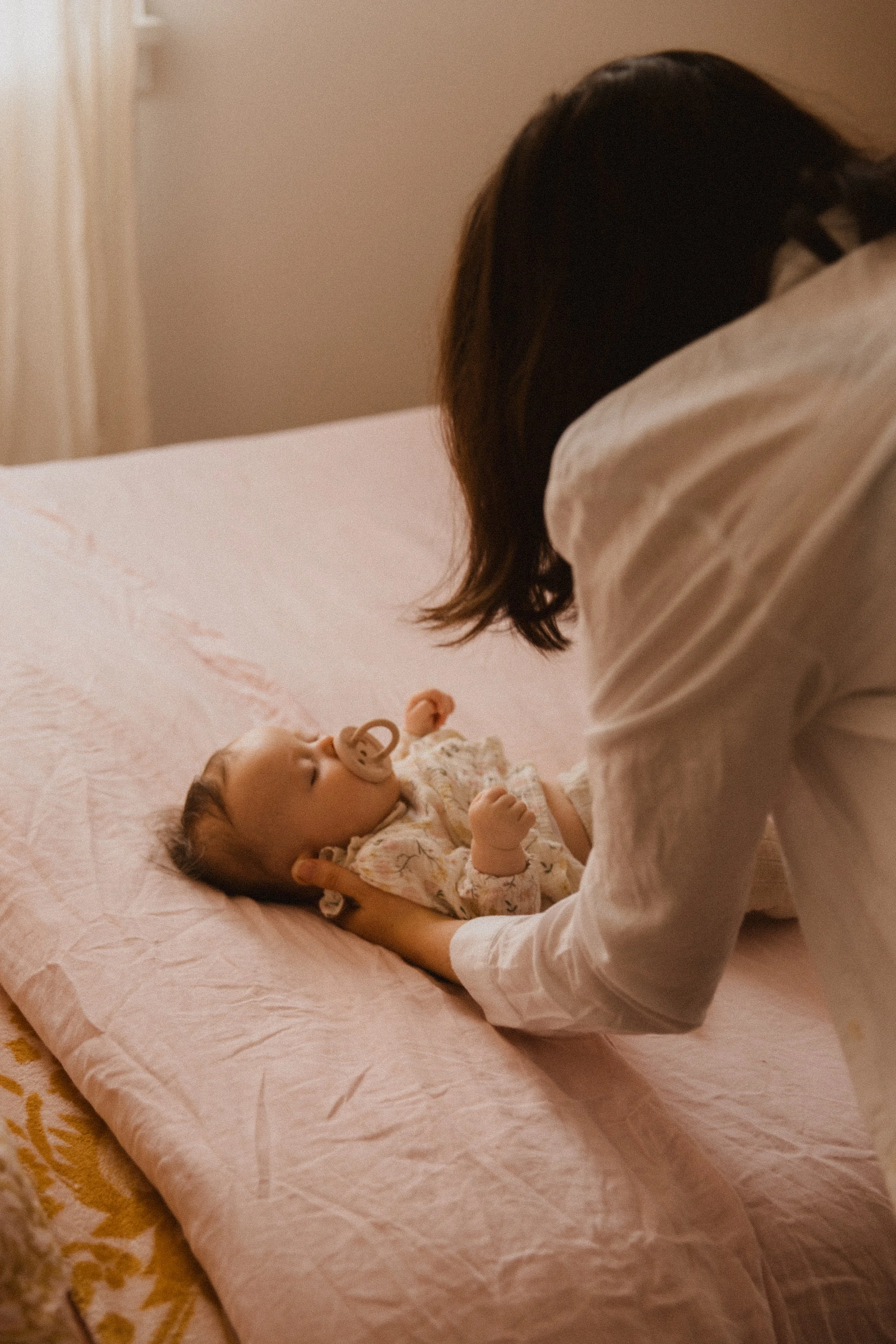 A woman gently holding a sleeping baby with a pacifier, lying on a pink blanket on a bed in a softly lit room.