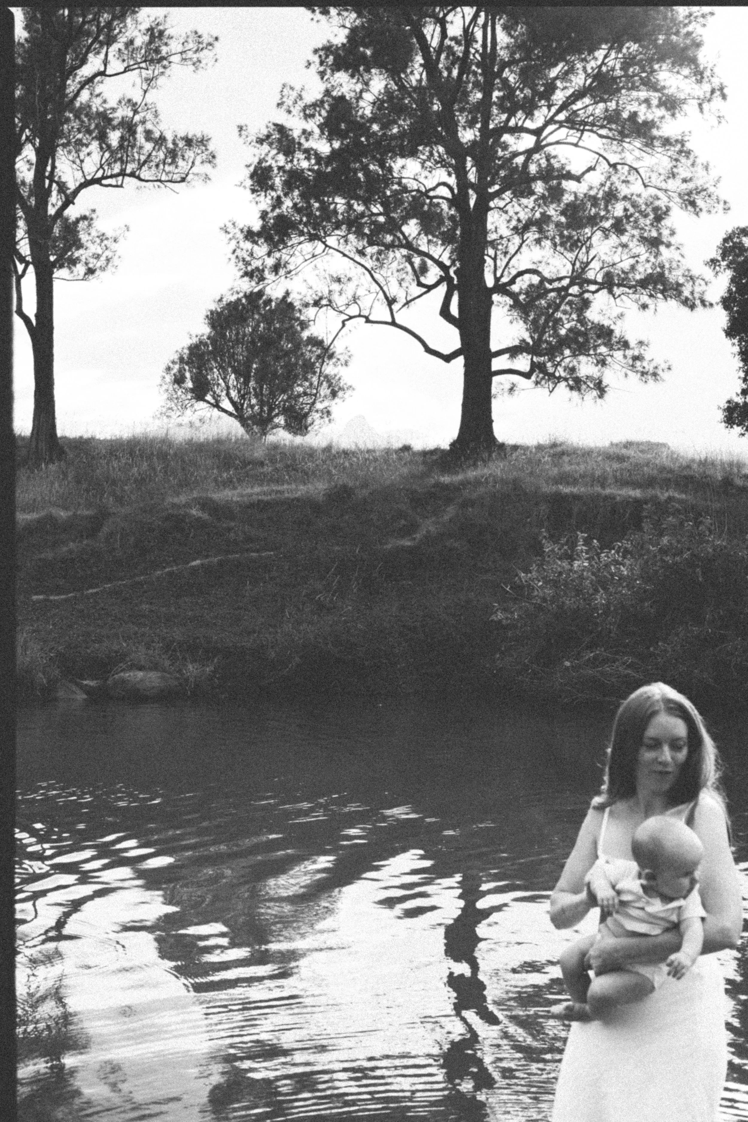 A woman holding a baby near a body of water with trees and grass in the background, in black and white.
