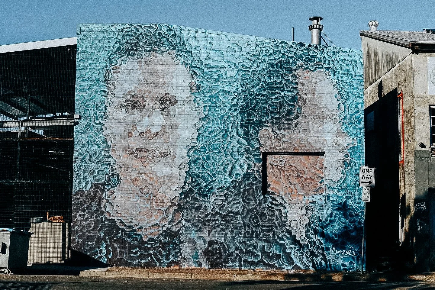 A large mural on a brick wall depicts two women's faces; the left woman has curly hair, prominent lips, and a serious expression, and the right woman has an afro hairstyle with a similar serious expression, with artistic textured shading in shades of