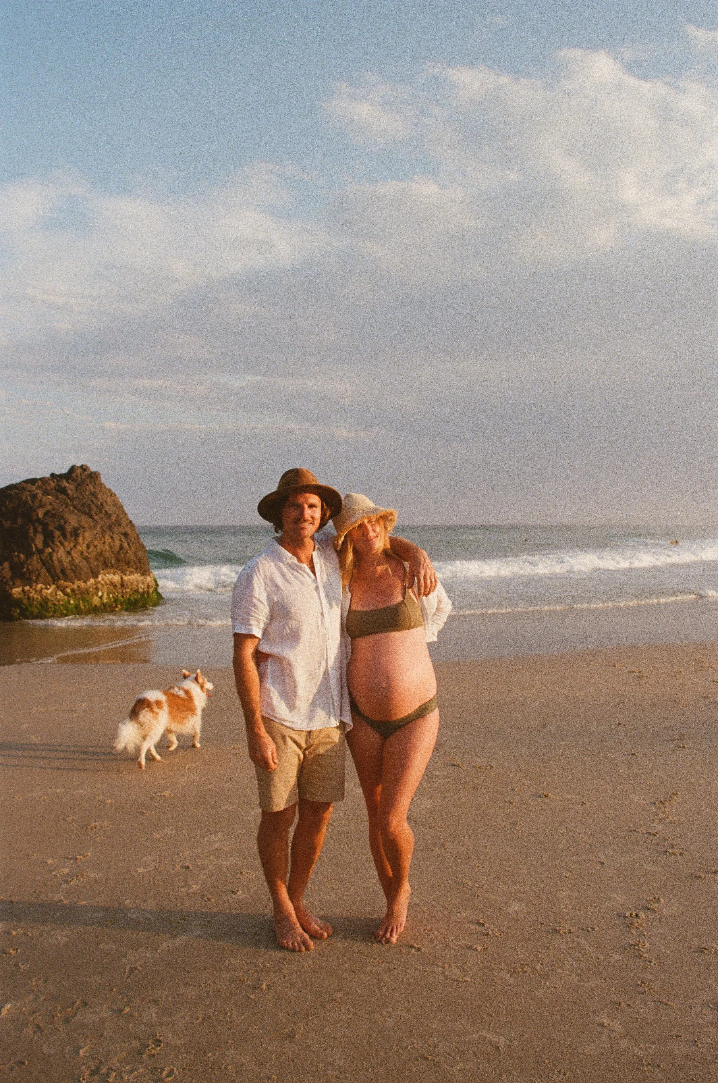 A couple and a dog on the beach, with the ocean and cloudy sky in the background. The woman is pregnant and wearing a swimsuit and sunhat, while the man is dressed in casual summer clothes and a wide-brimmed hat. The dog is white with brown markings