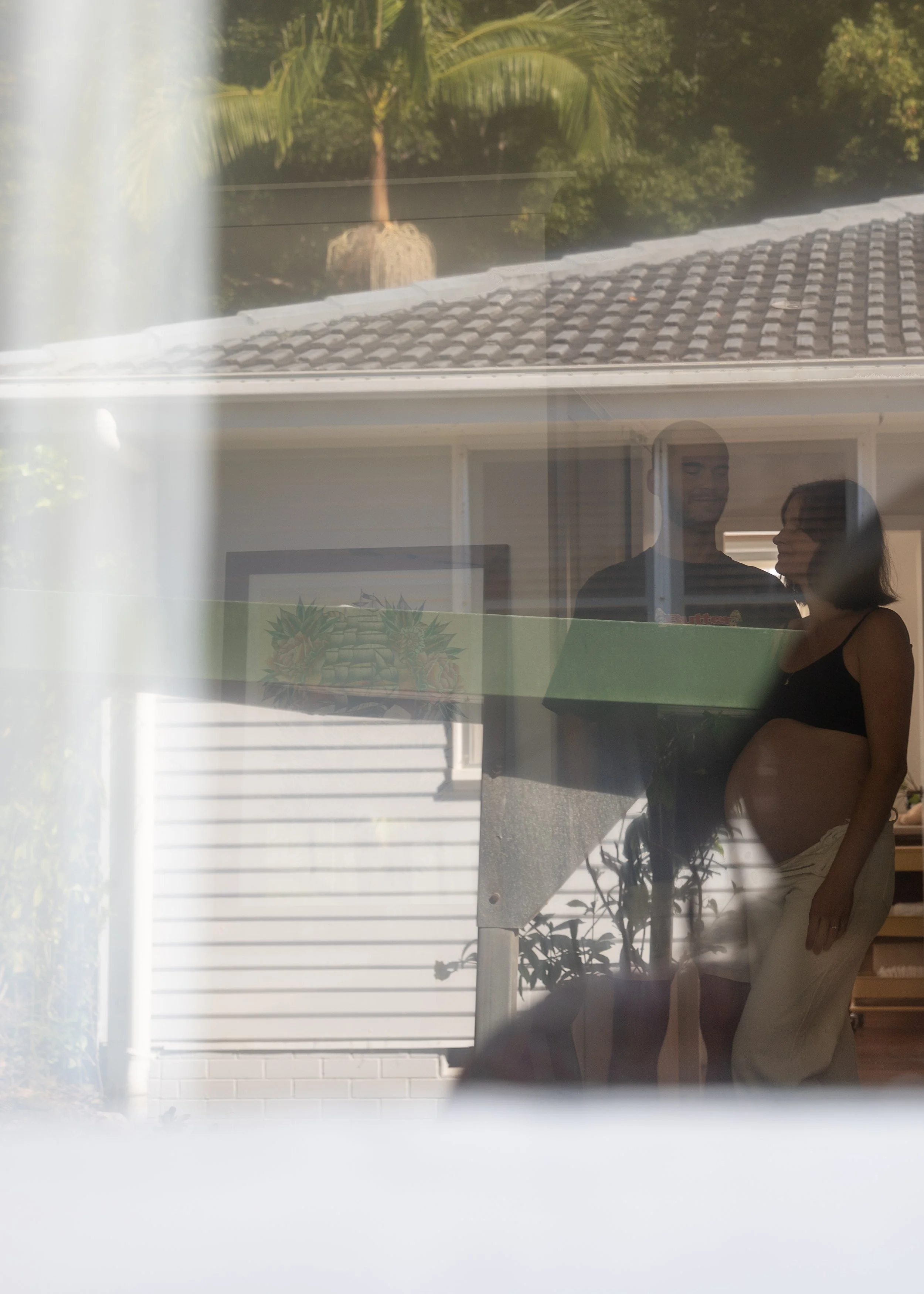 Reflection of a pregnant woman and a man with a cake inside a house, viewed through a window with curtains, with trees outside.