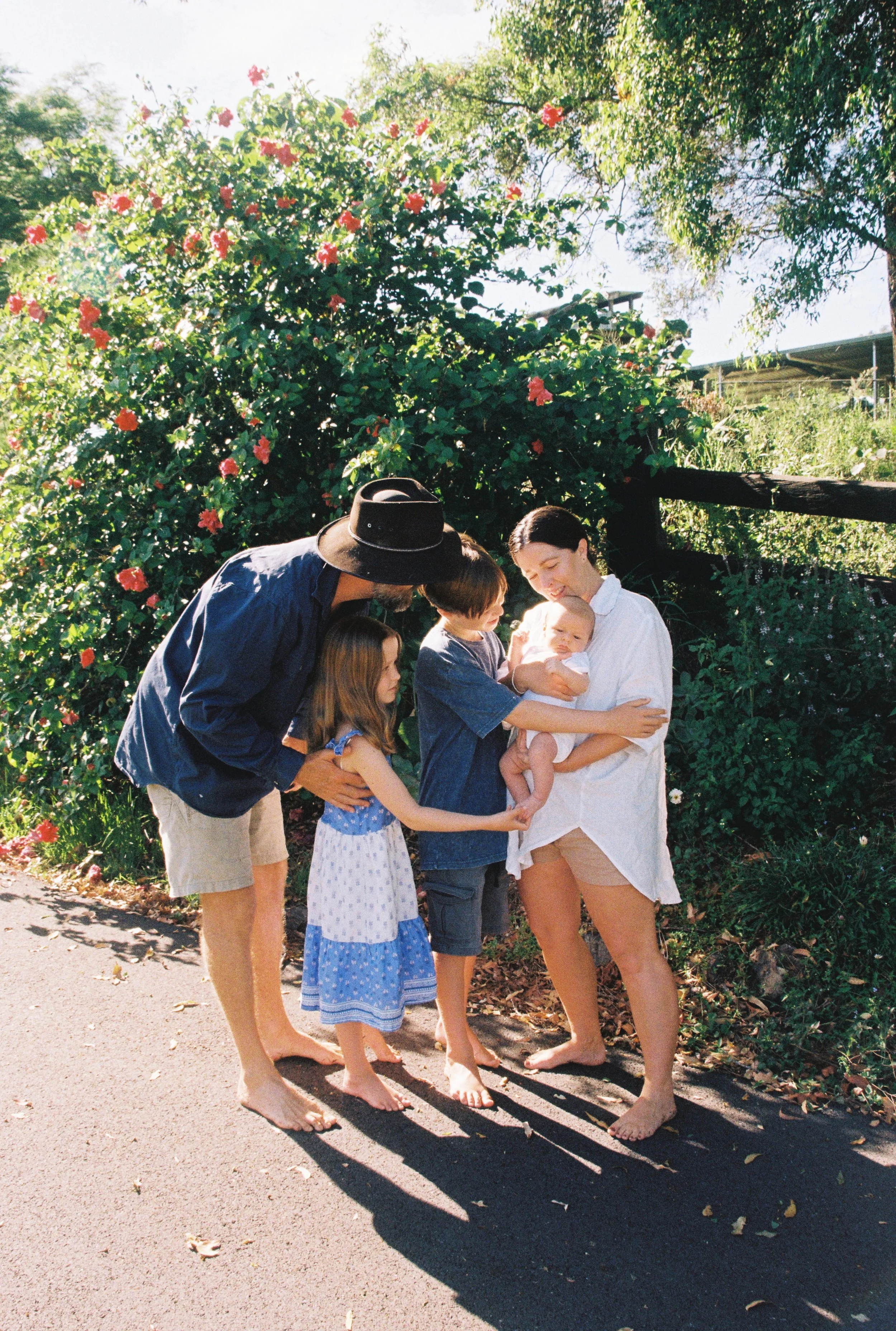 A family of five with two children and a baby, standing on a sidewalk near greenery and pink flowers, smiling and interacting outside on a sunny day.