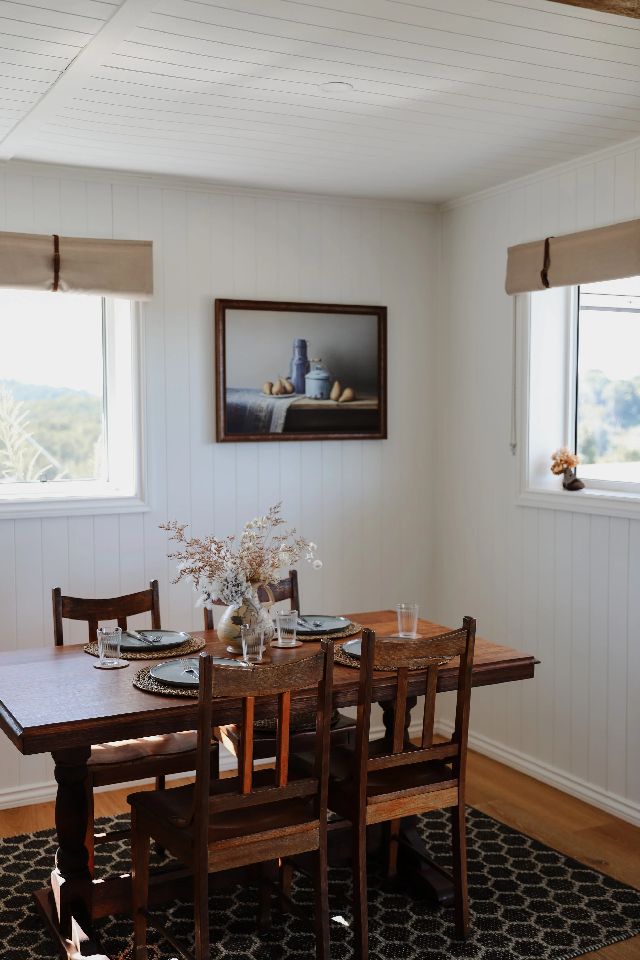 A cozy dining room with a wooden table set for four, featuring plates, glasses, and silverware, with a decorative vase containing dried flowers in the center. The room has white paneled walls, two windows with beige shades, and a framed still life pa