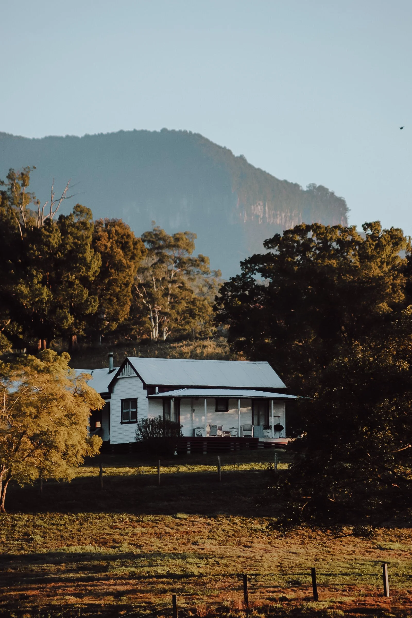 A white house with a metal roof surrounded by trees and a scenic mountain backdrop during sunset.