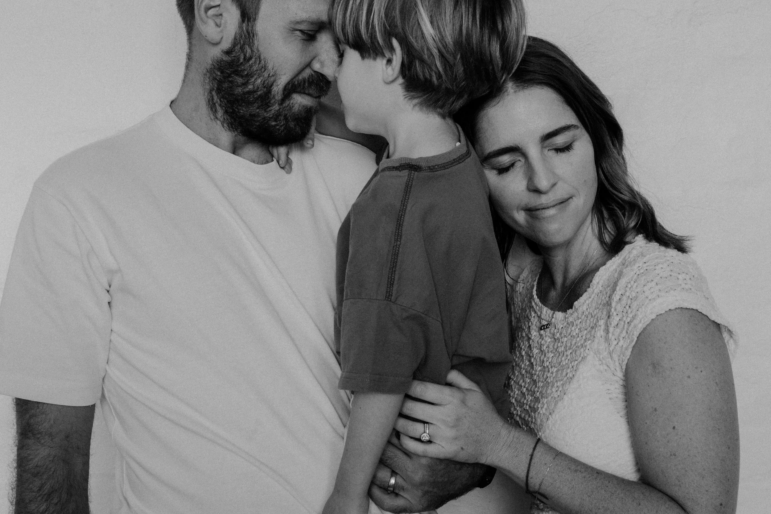 A black and white photo of a family of three, with a father, mother, and young son. The father has a beard, and the mother has dark hair. They are holding the son close, showing affection and warmth.