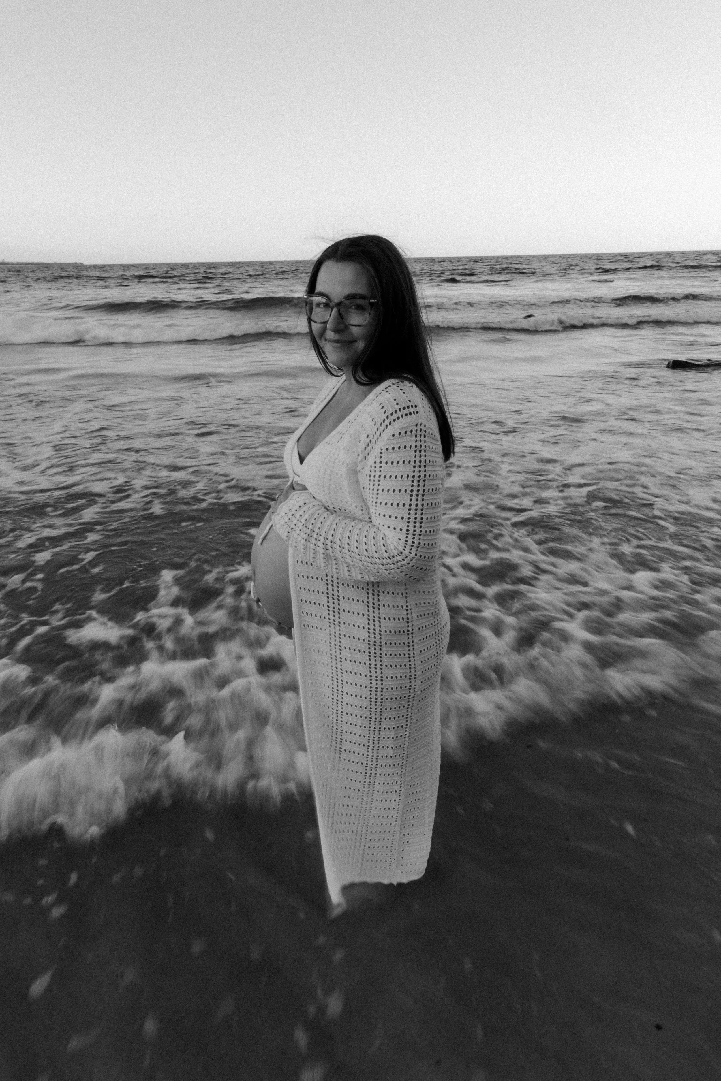 A pregnant woman in glasses standing in the ocean waves at the beach, wearing a white crochet cover-up and smiling.