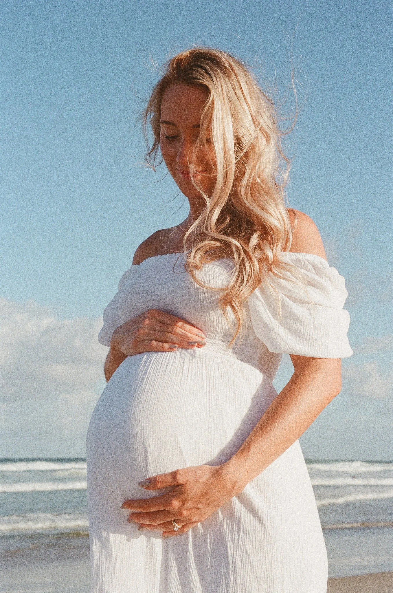 Pregnant woman with long, wavy blonde hair standing on the beach, wearing a white off-the-shoulder dress, gently holding her baby bump.