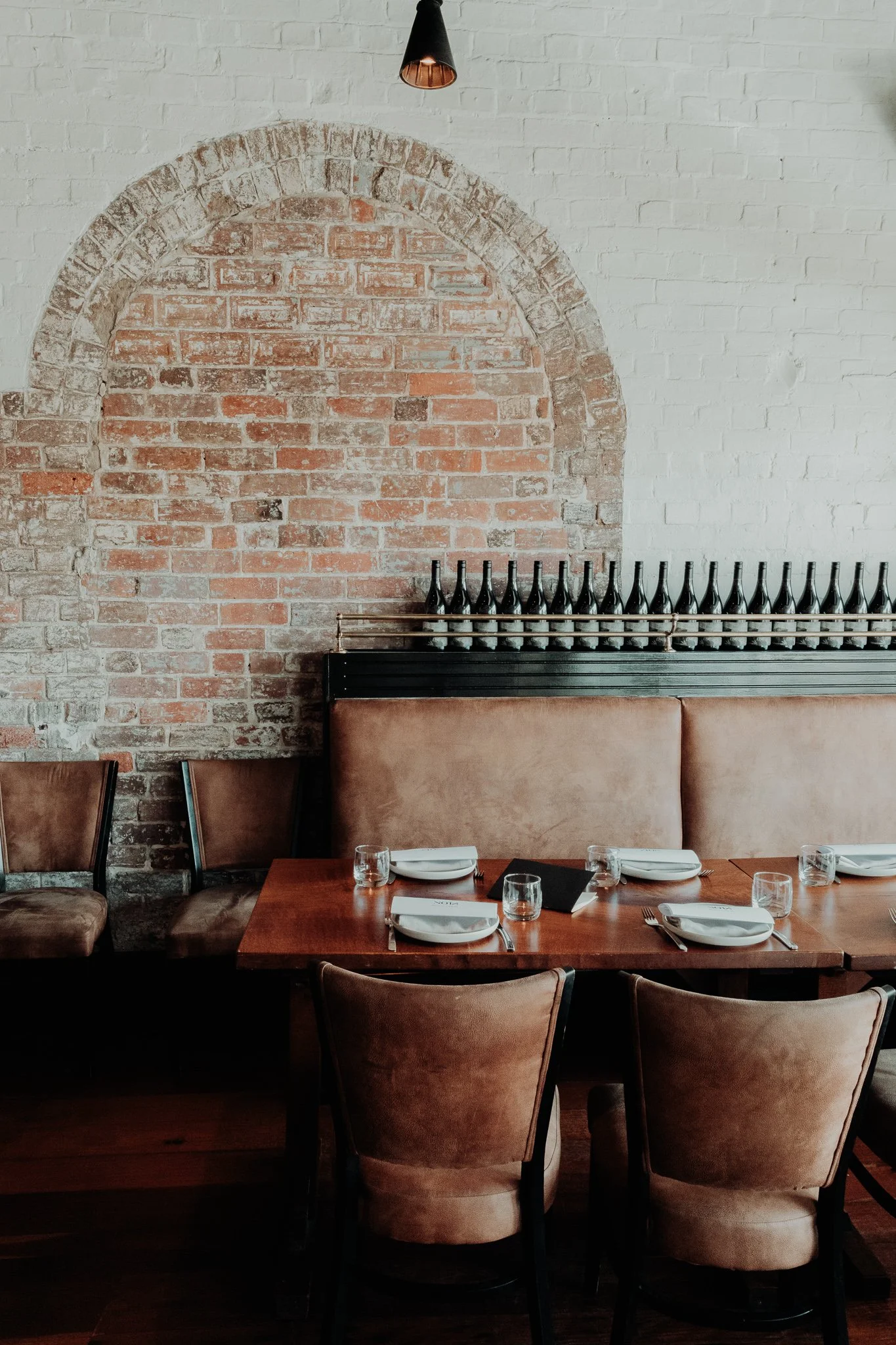 A cozy restaurant interior with a brick wall, a row of empty wine bottles on a shelf, a wooden table set with plates, glasses, and cutlery, and upholstered chairs.