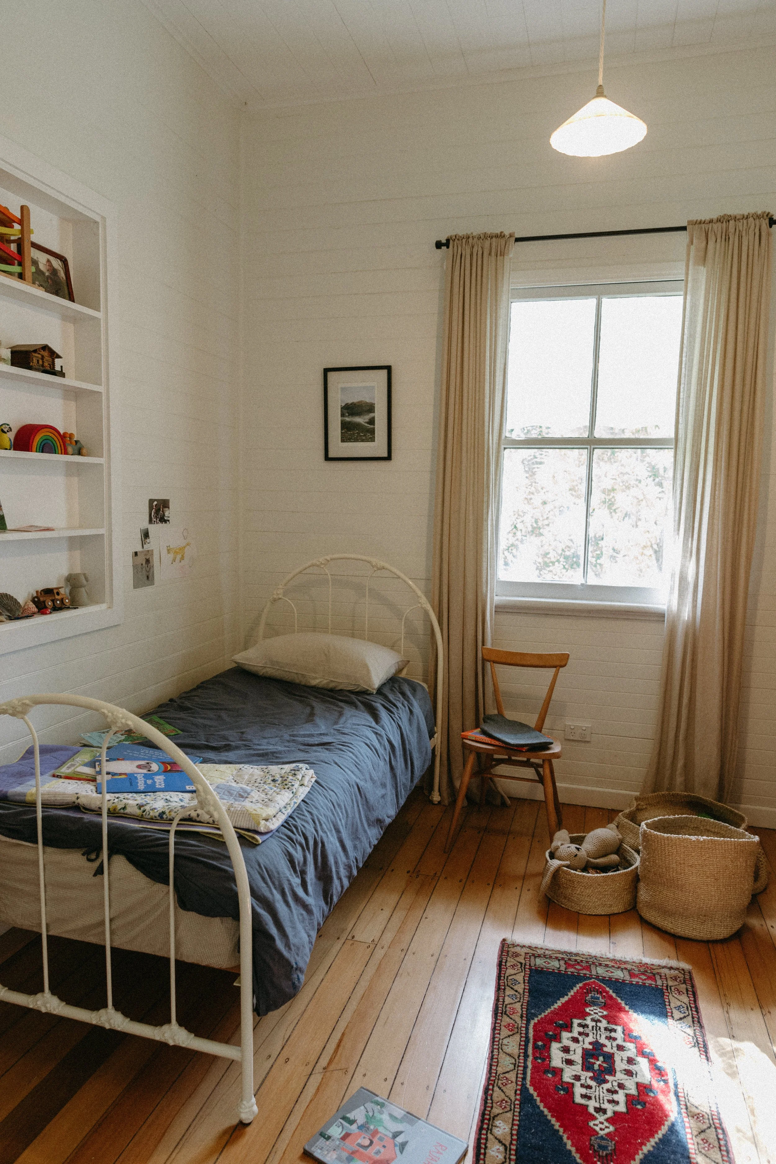 A cozy bedroom with a white metal bed, beige curtains, a wooden chair, and a small rug on the wooden floor. There are baskets and plush toys near the window, and books and framed pictures on the walls and shelves.