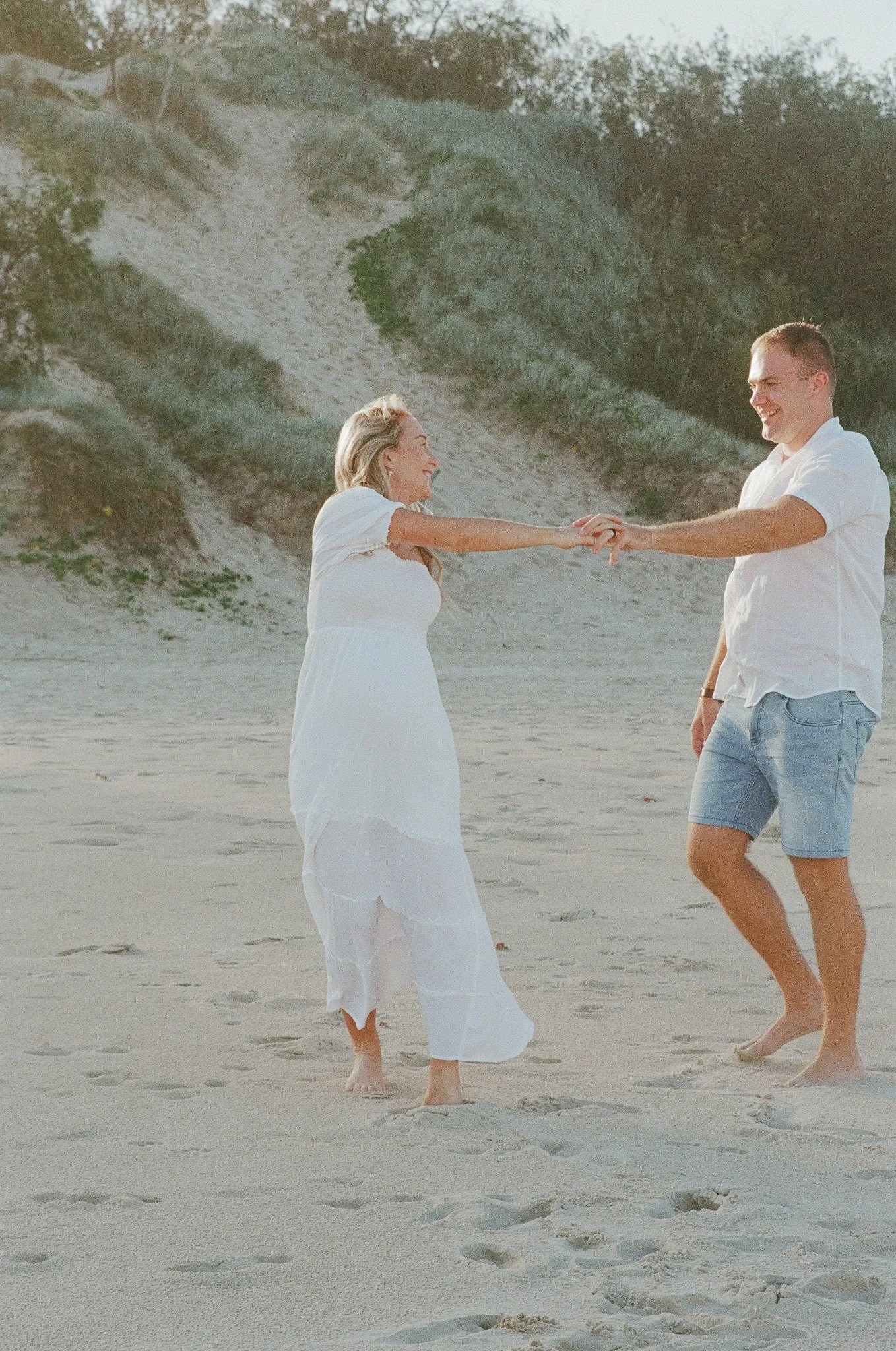 A couple on a beach holding hands and smiling at each other, with dunes and trees in the background.