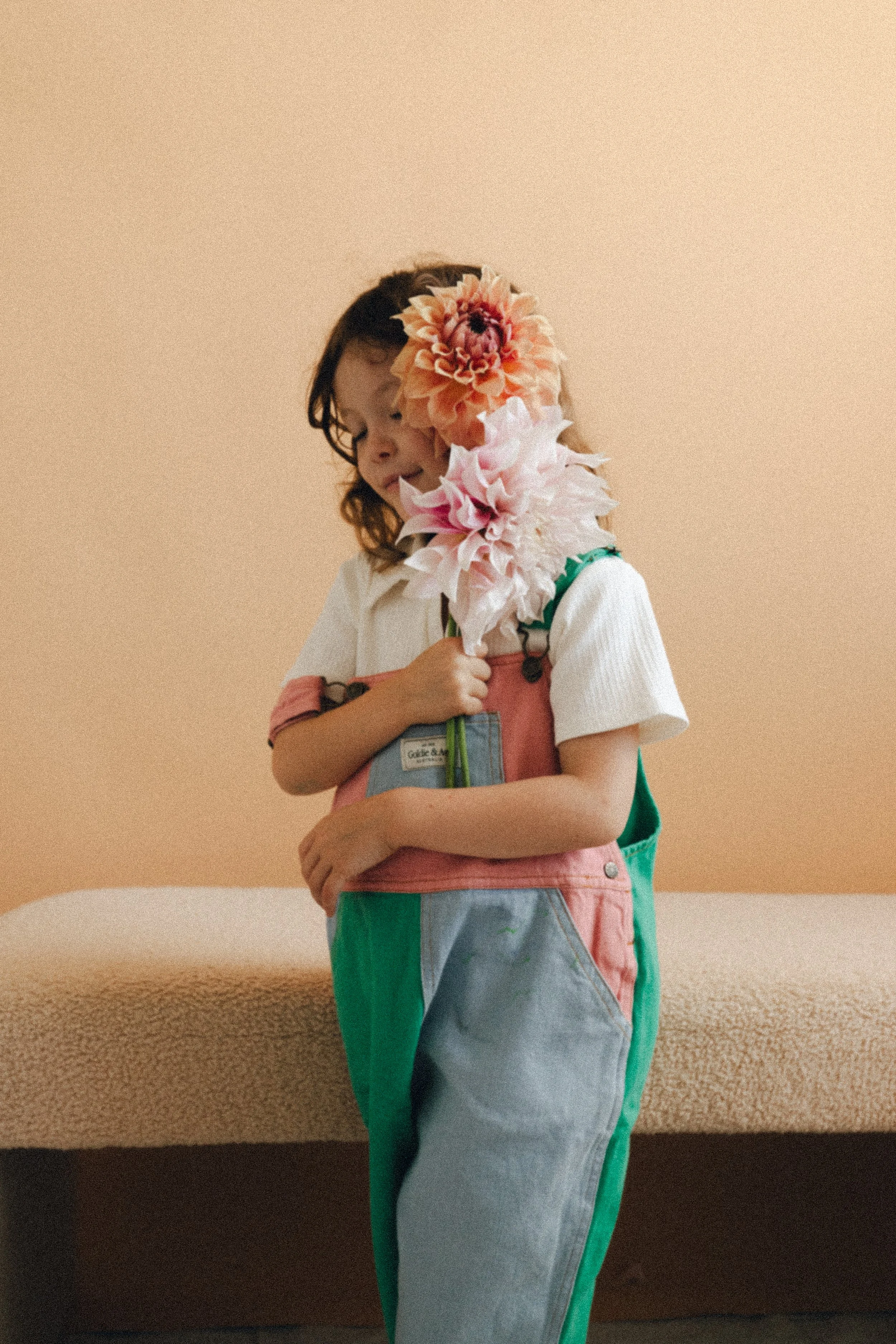 A girl holding two large artificial flowers, one pink and one peach, standing in front of a beige background with a beige couch behind her.