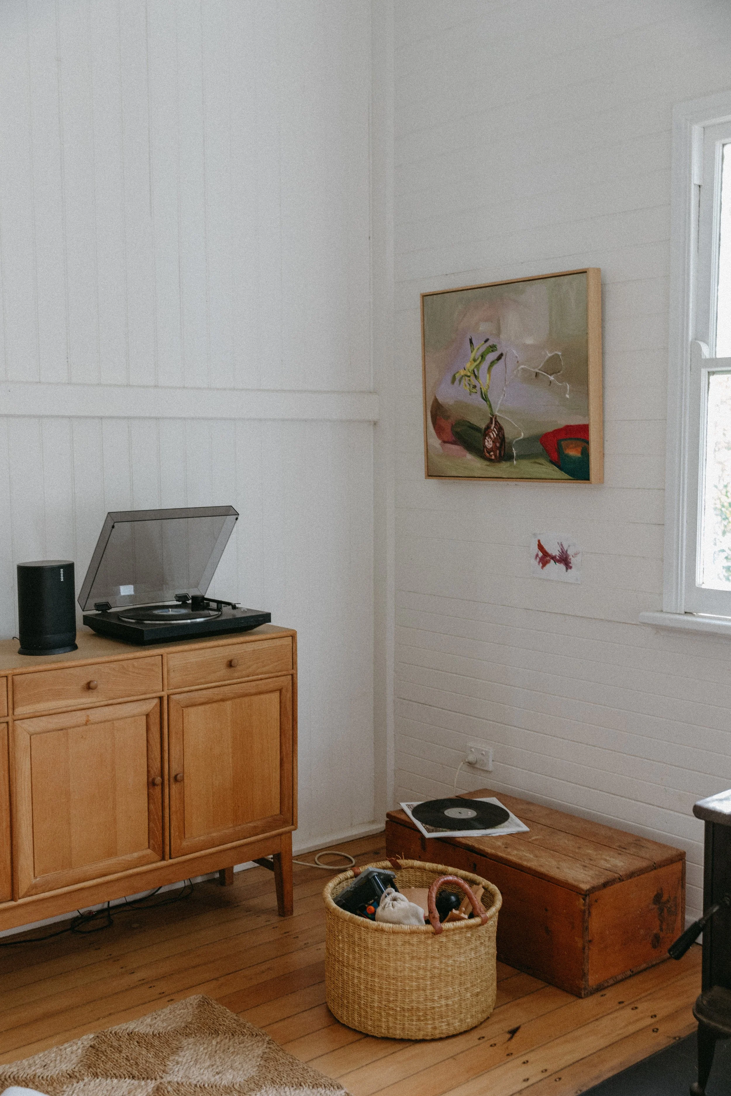 A corner of a room with a wooden cabinet holding a record player and speaker, a wicker basket with toys on the floor, a wooden chest, a framed painting, a small paper with a lobster drawing, and a window with sunlight streaming in.
