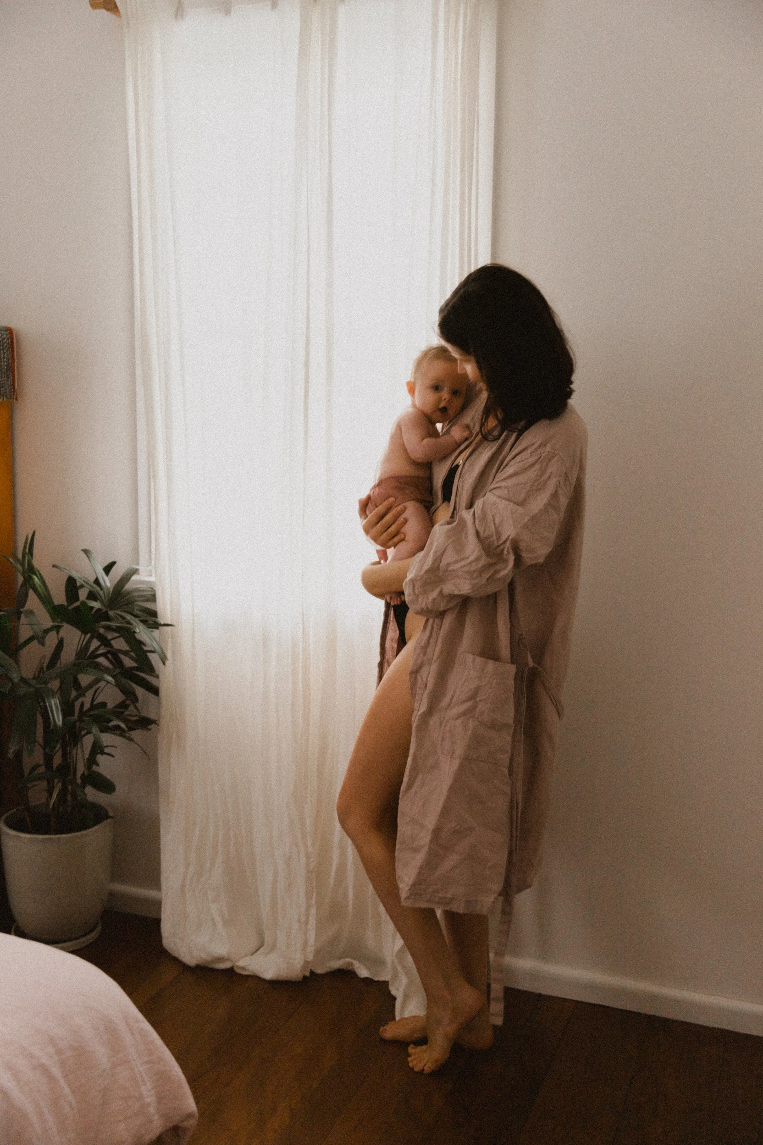 A woman holding a small baby in a softly lit room with white curtains and a potted plant.