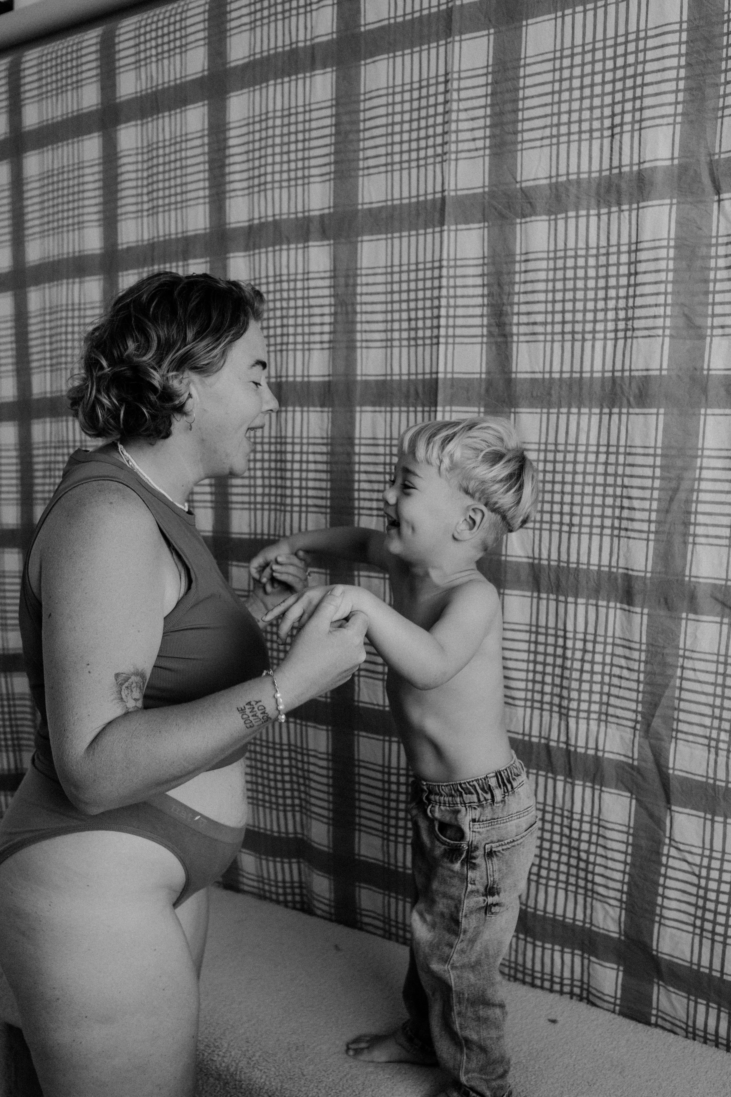 A woman and a young boy are smiling and playing together indoors against a plaid curtain backdrop. The woman is wearing a tank top and underwear, and the boy is shirtless in jeans. They are holding hands and looking at each other happily.