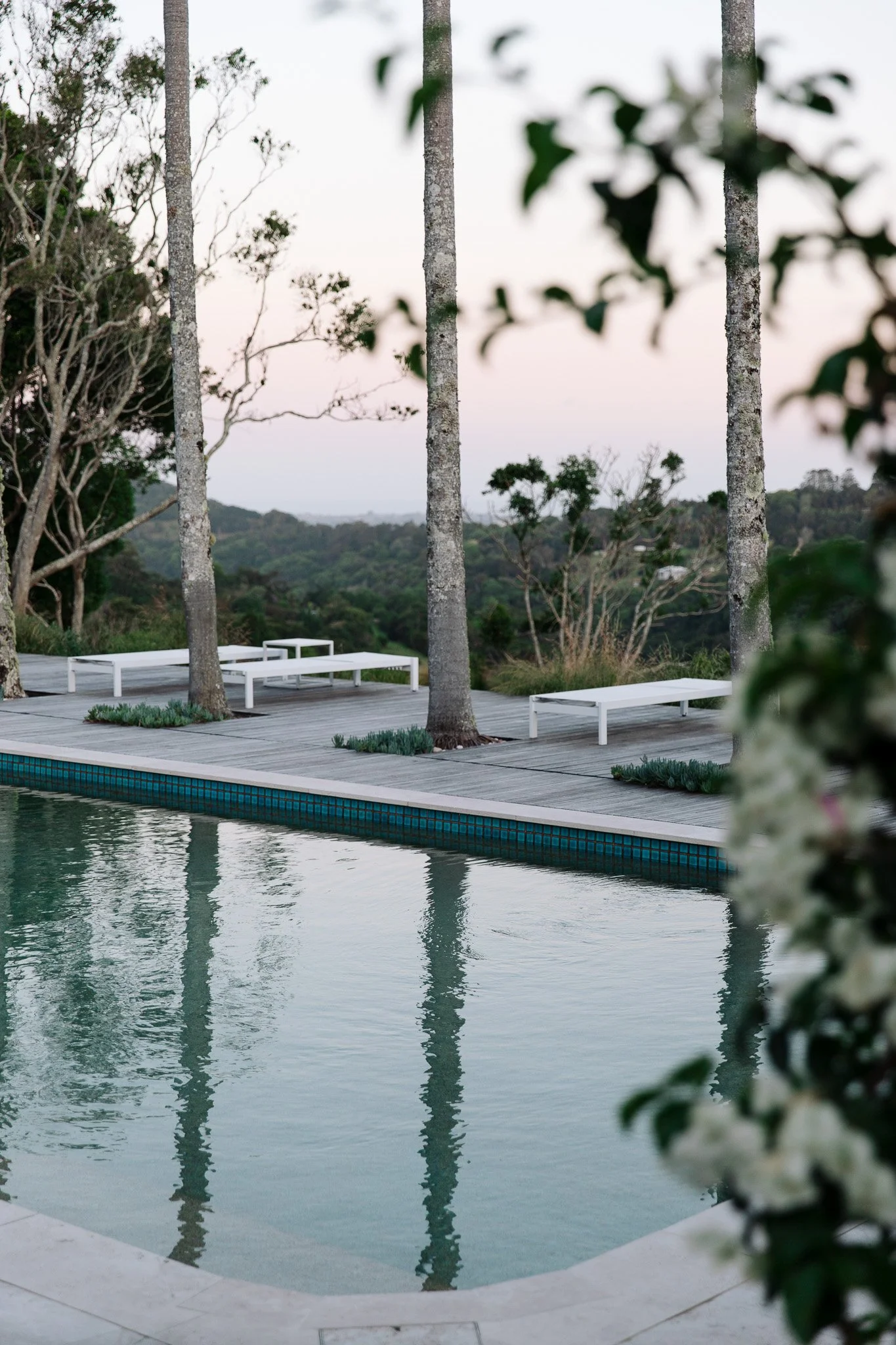 A view of a swimming pool with a wooden deck, surrounded by trees and greenery, with lounge chairs on the deck and a scenic landscape in the background during dusk or early evening.